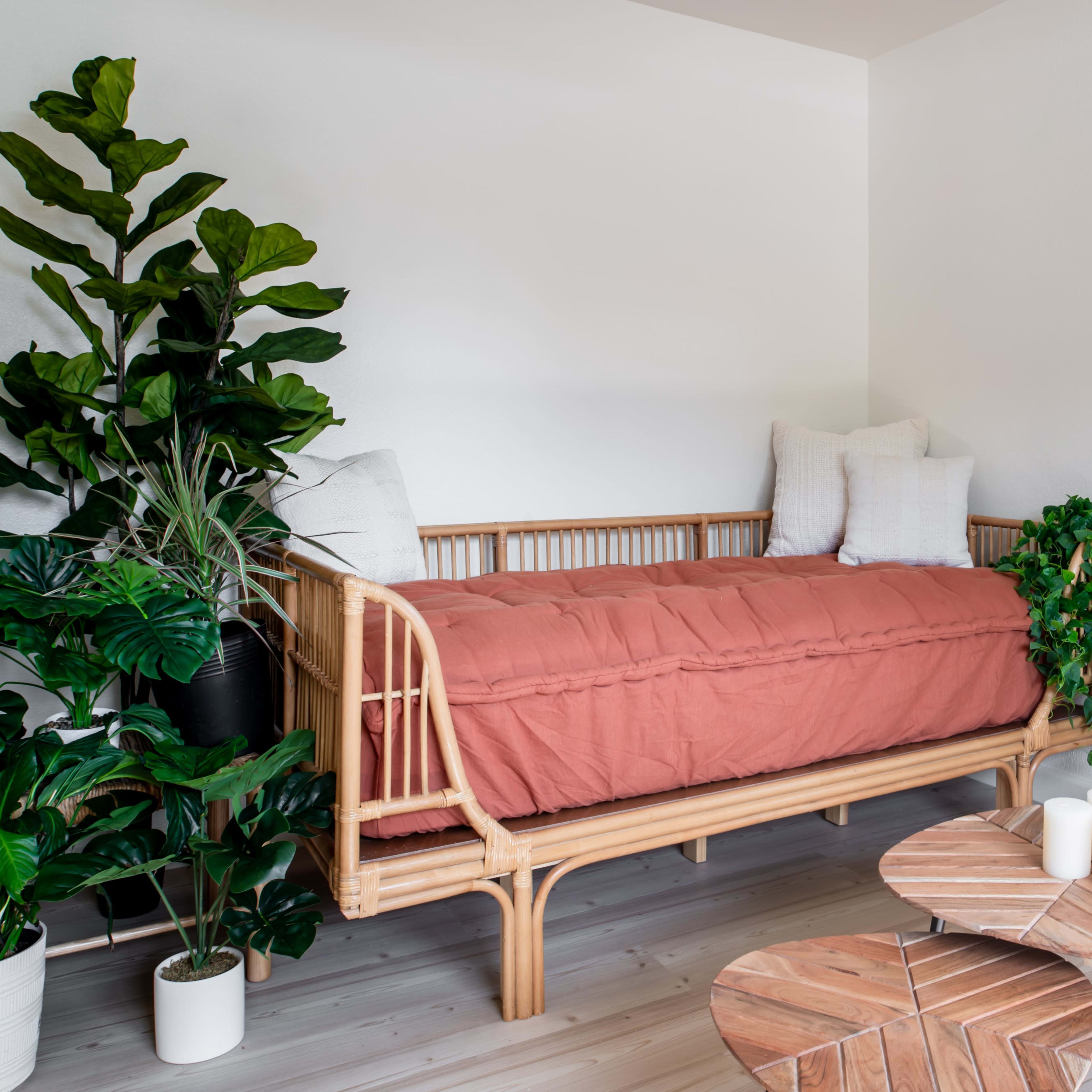 A rattan daybed with an orange cover is positioned against a white wall, surrounded by green plants and a small wooden table set.