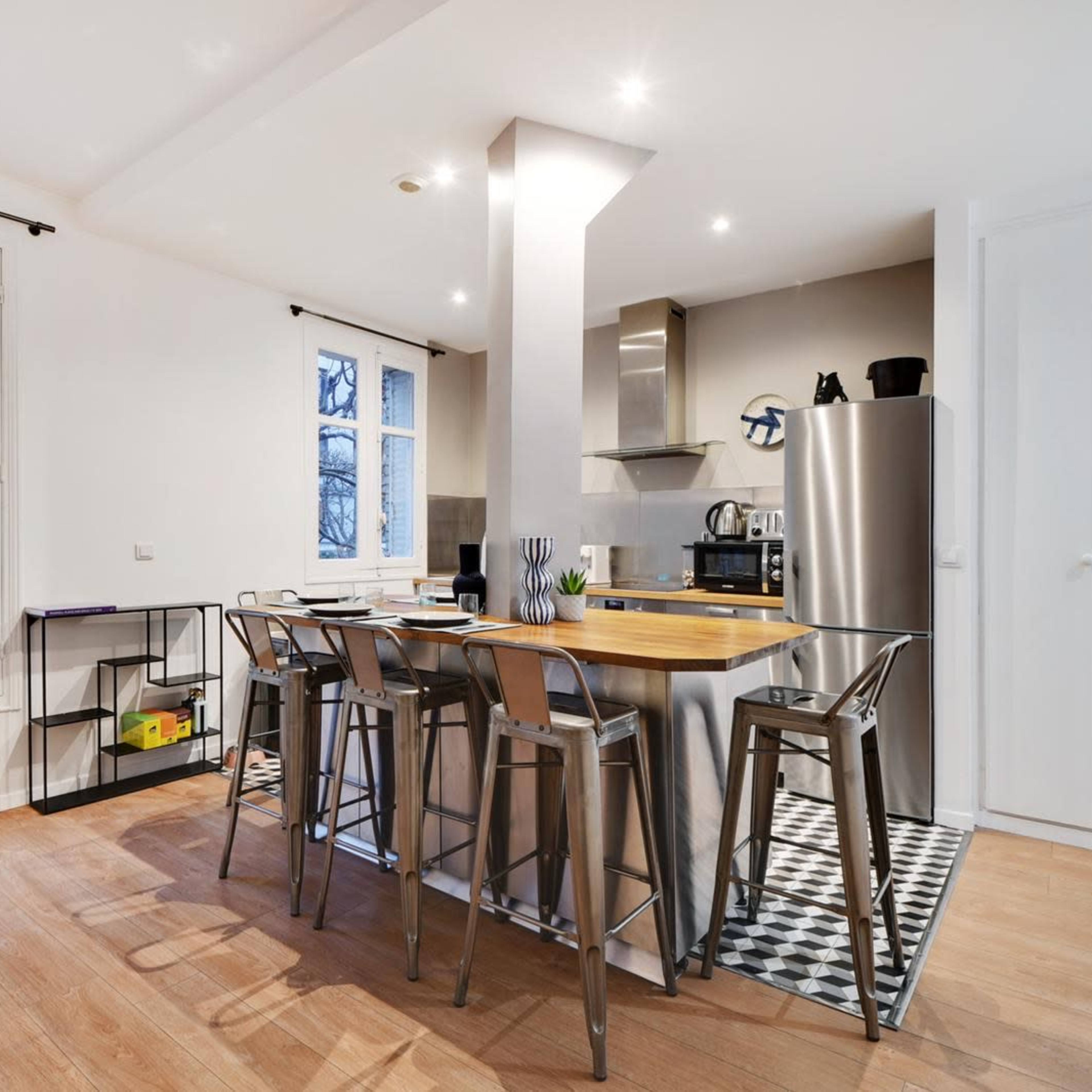 A modern kitchen with a central island, high stools, stainless steel appliances, and large windows overlooking a balcony.