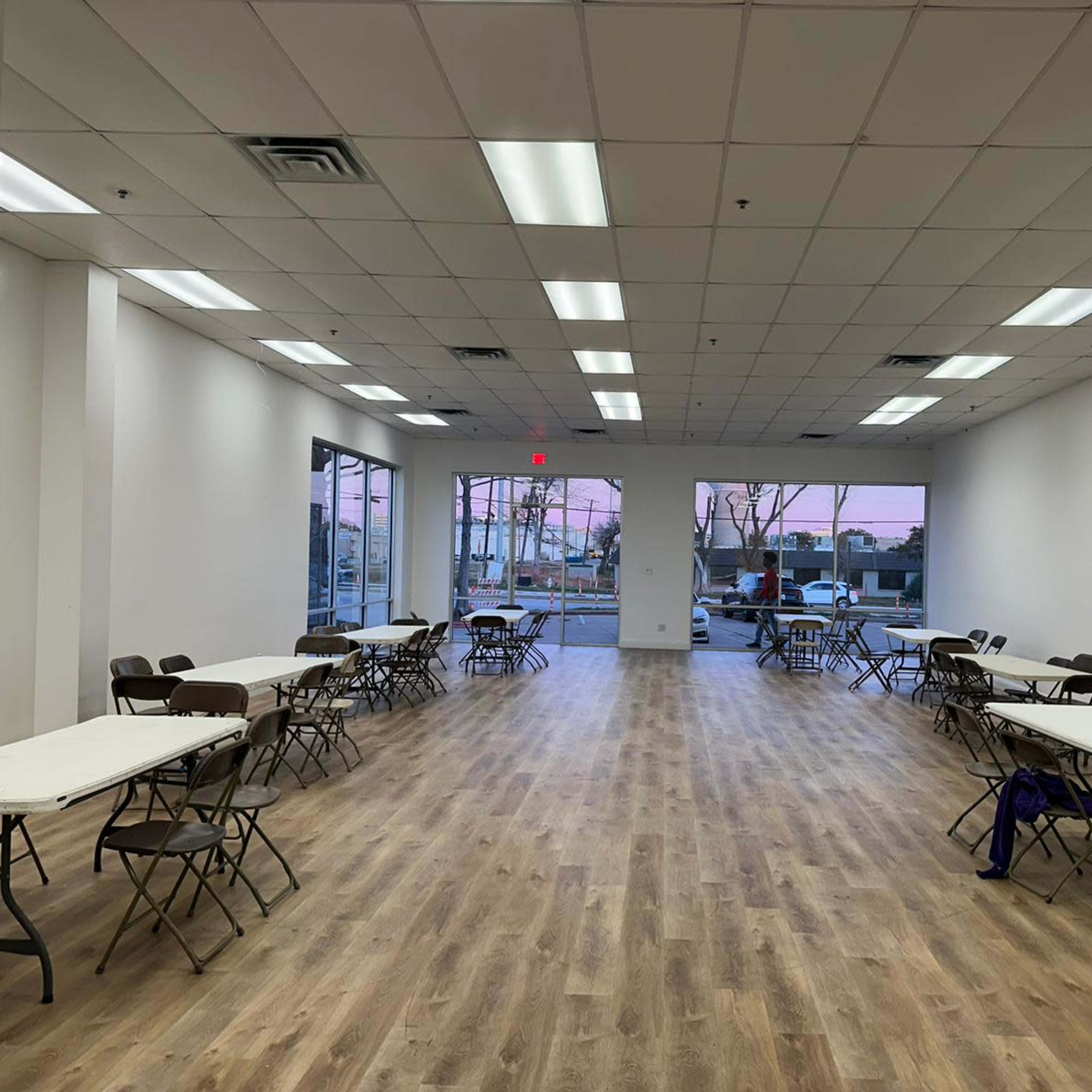 A large, empty room with multiple tables and chairs arranged in rows, large windows on one side, and a pale wooden floor.