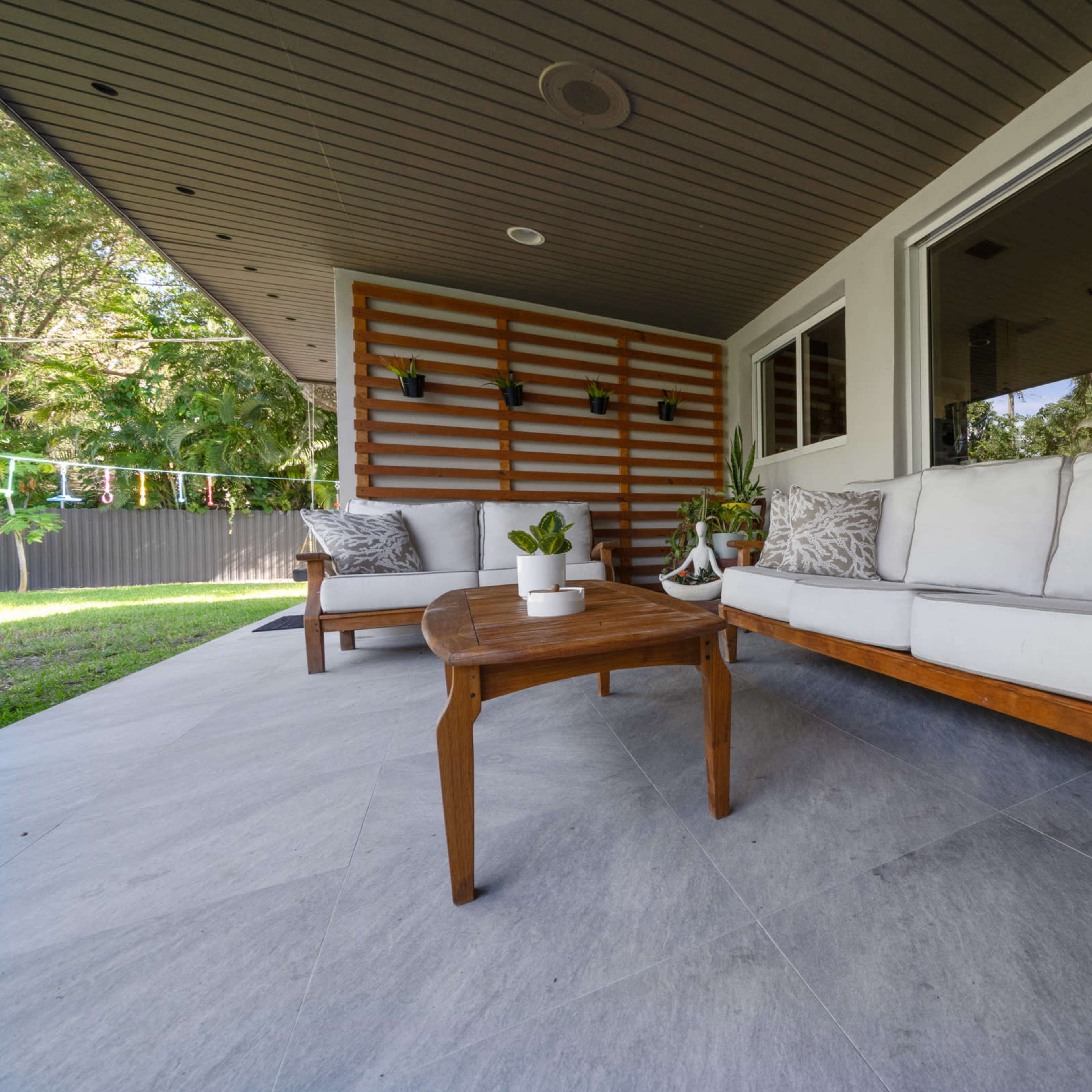 A covered patio features a wooden coffee table and a light-colored sofa with cushions, surrounded by greenery.