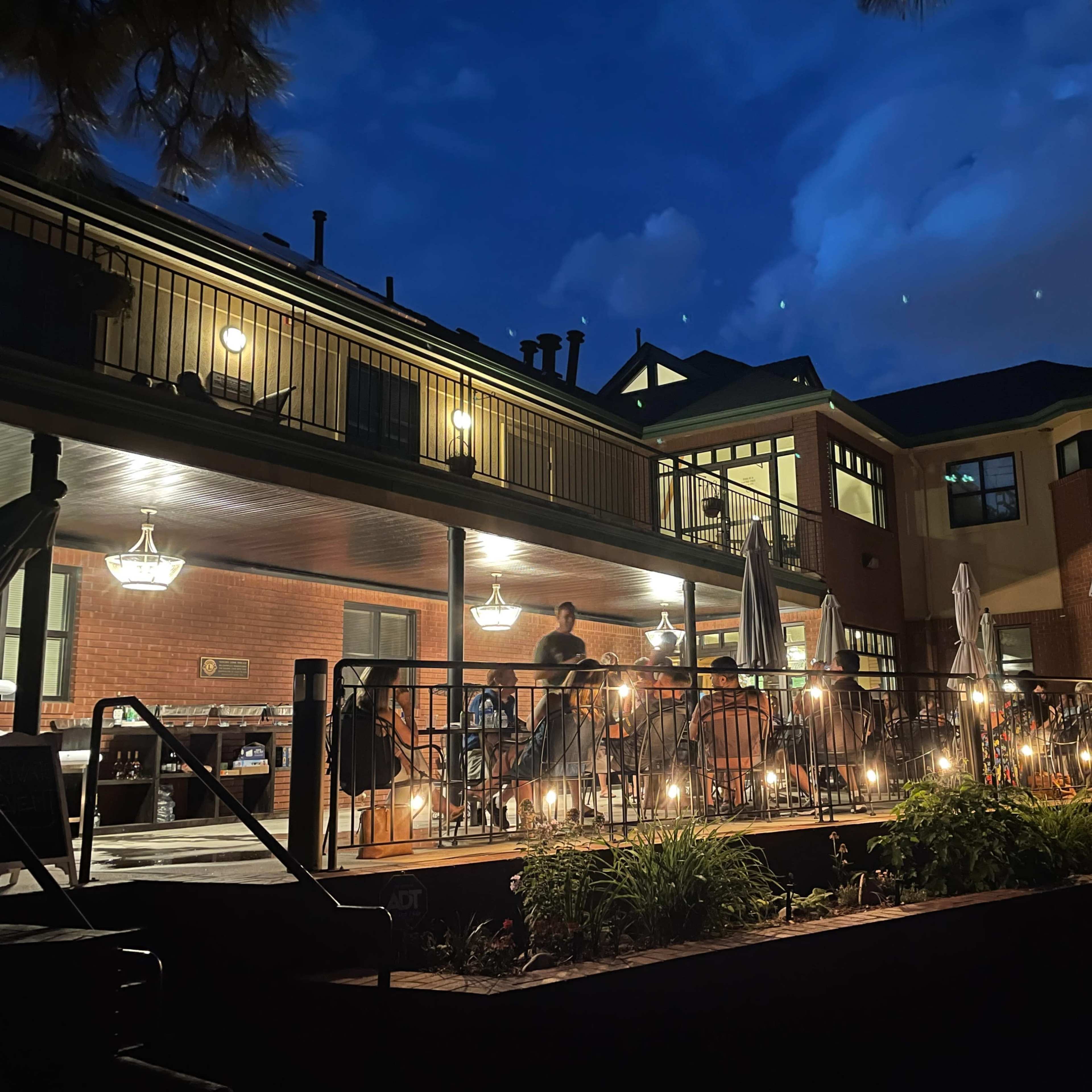 A well-lit patio with string lights and several people dining is attached to a brick building under a night sky.