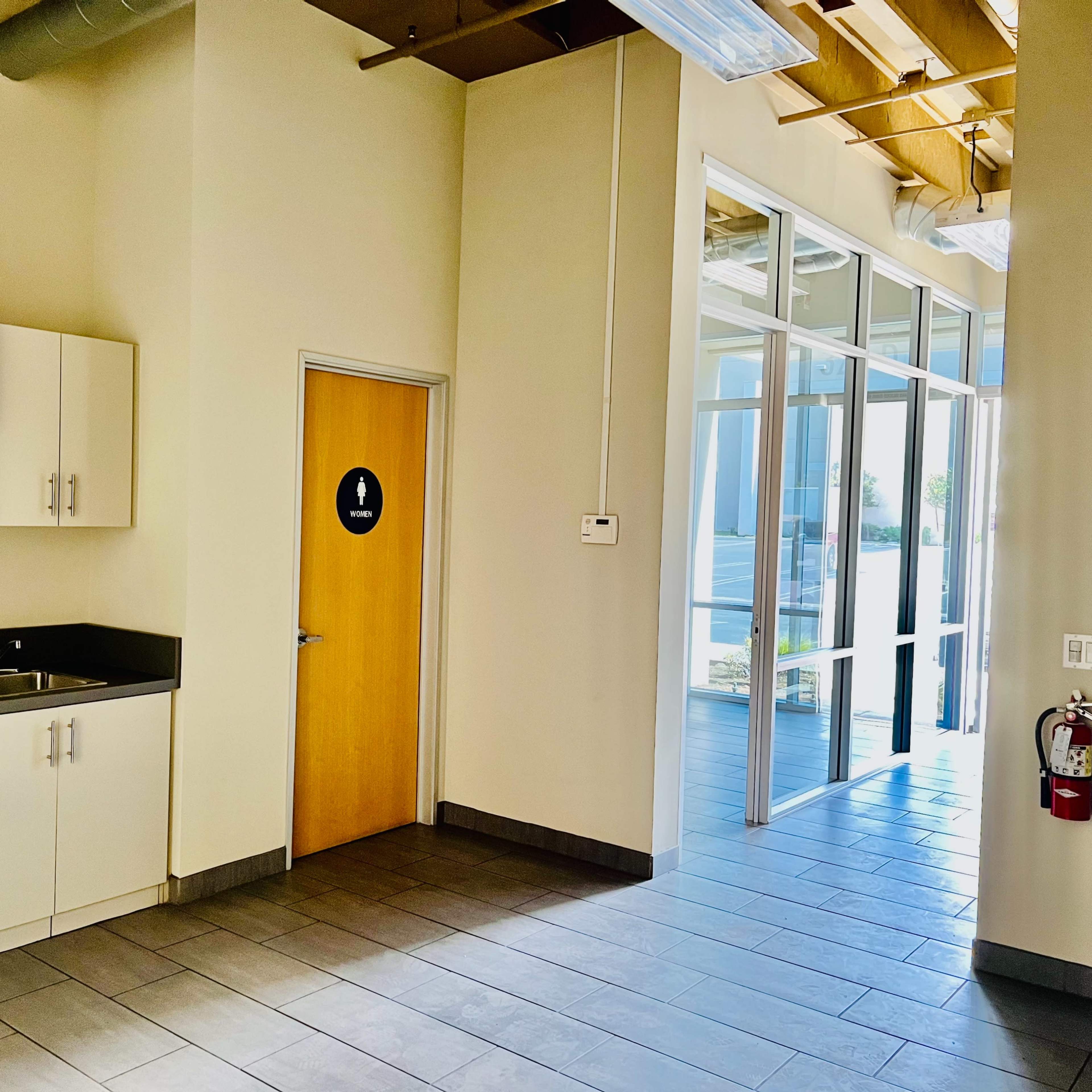 A modern interior space featuring a small kitchenette on one side and a restroom door on the other, with large windows allowing natural light.