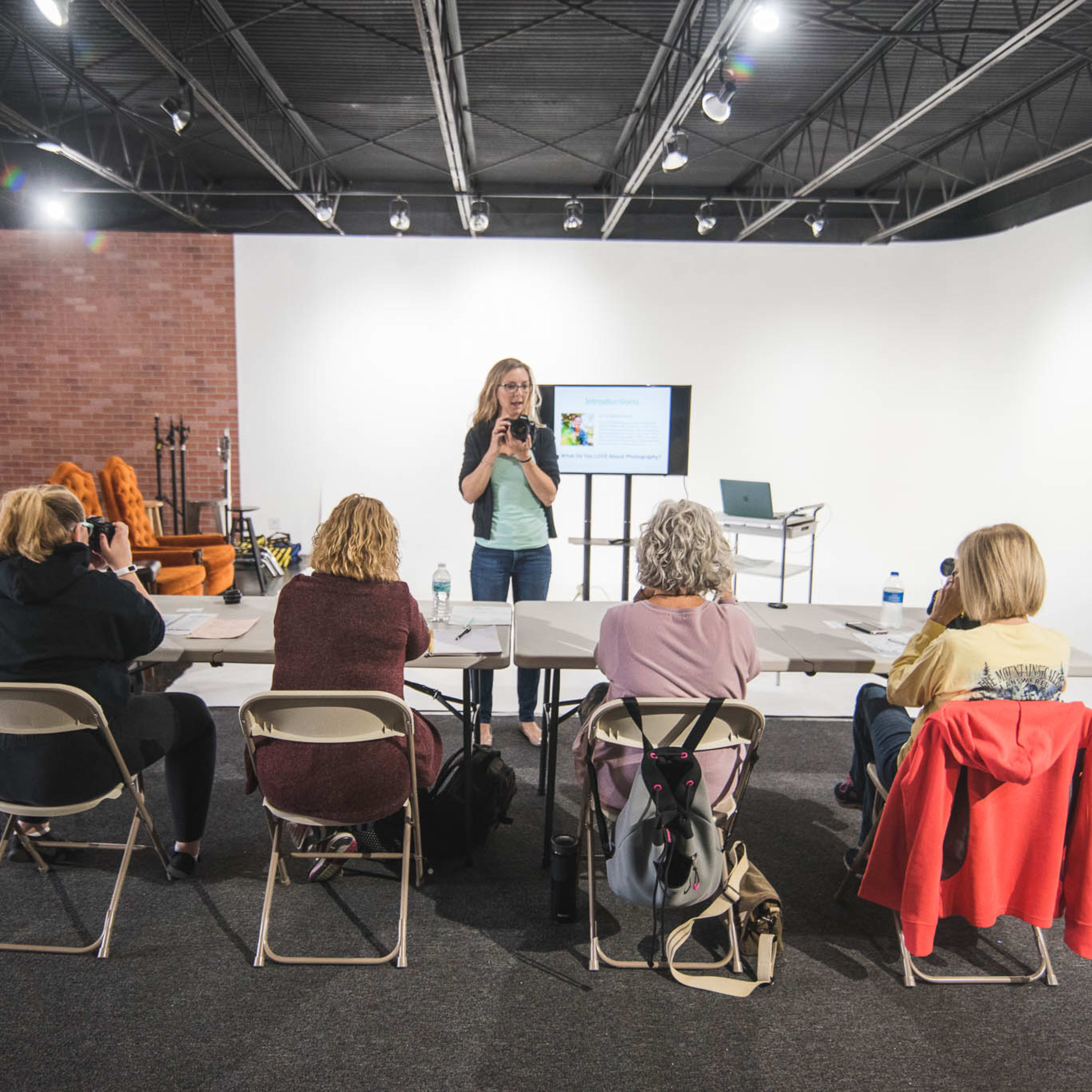 A woman stands at the front of a brightly lit room, presenting to a group of four seated individuals at a table.