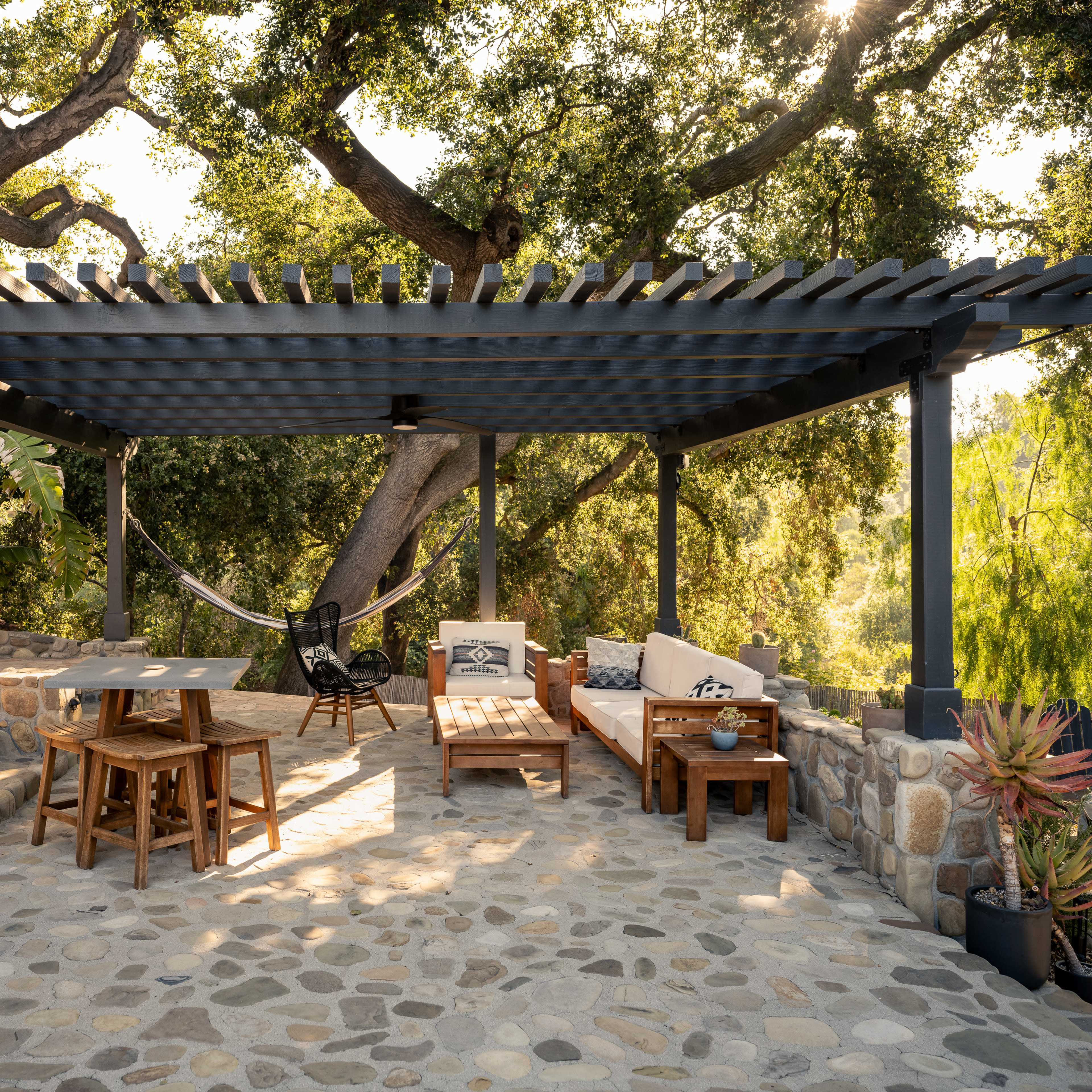A shaded outdoor seating area features wooden furniture, a hammock, and natural stone flooring under a pergola surrounded by trees.