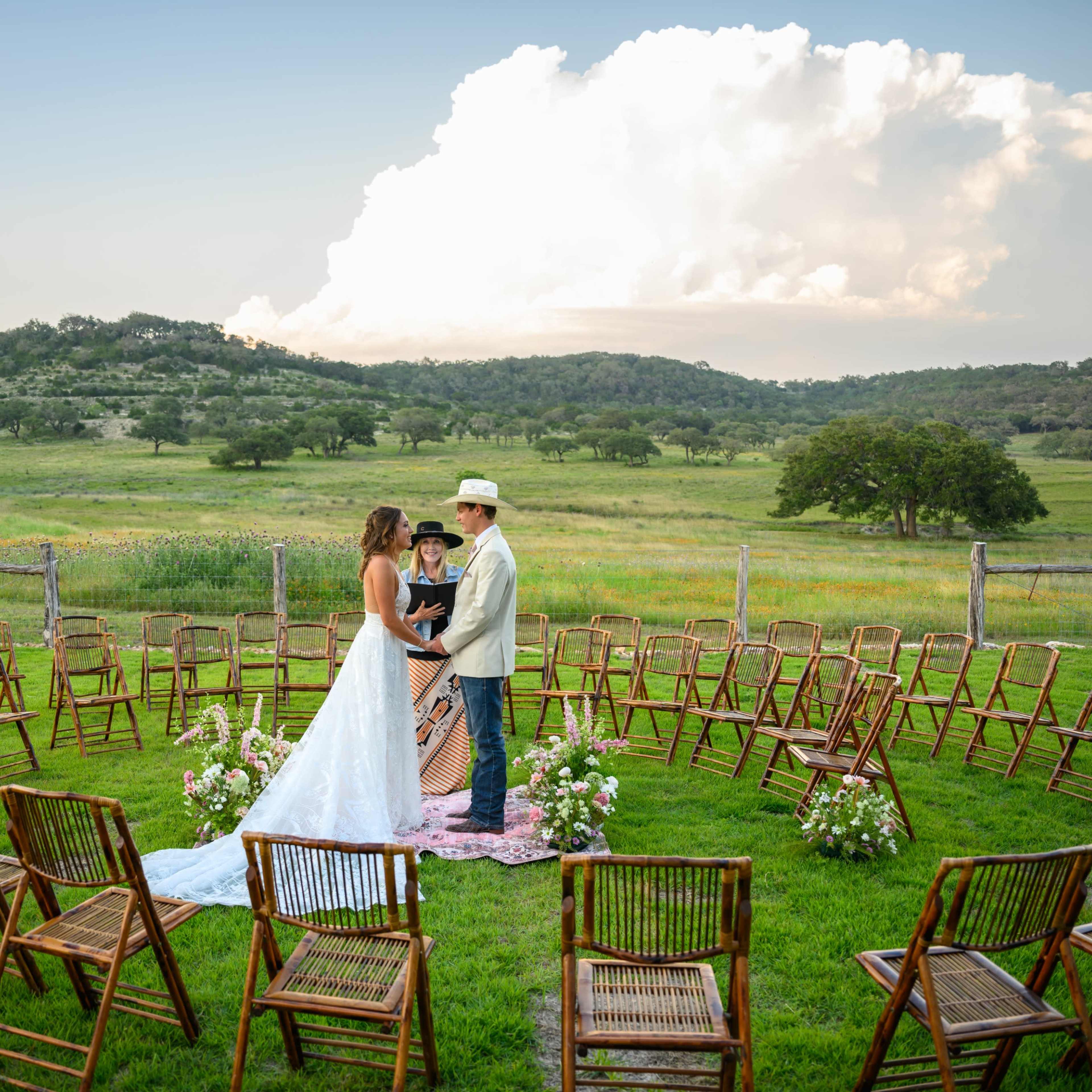 A couple stands in a grassy clearing surrounded by a circle of empty wooden chairs, with rolling hills and clouds in the background.