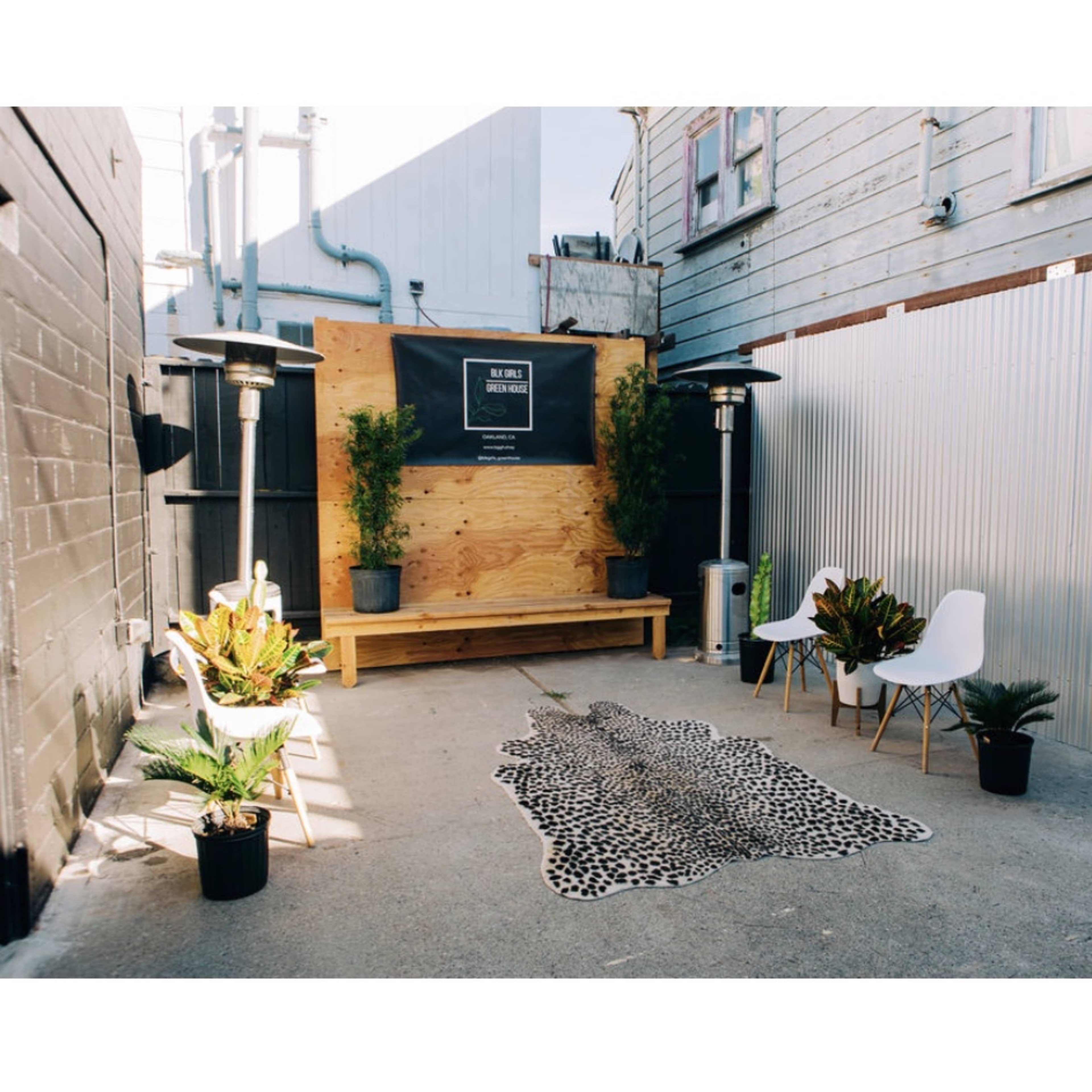 A small outdoor seating area with two white chairs, a wooden bench, potted plants, and a faux animal skin rug on a concrete floor, surrounded by walls of wood and metal.