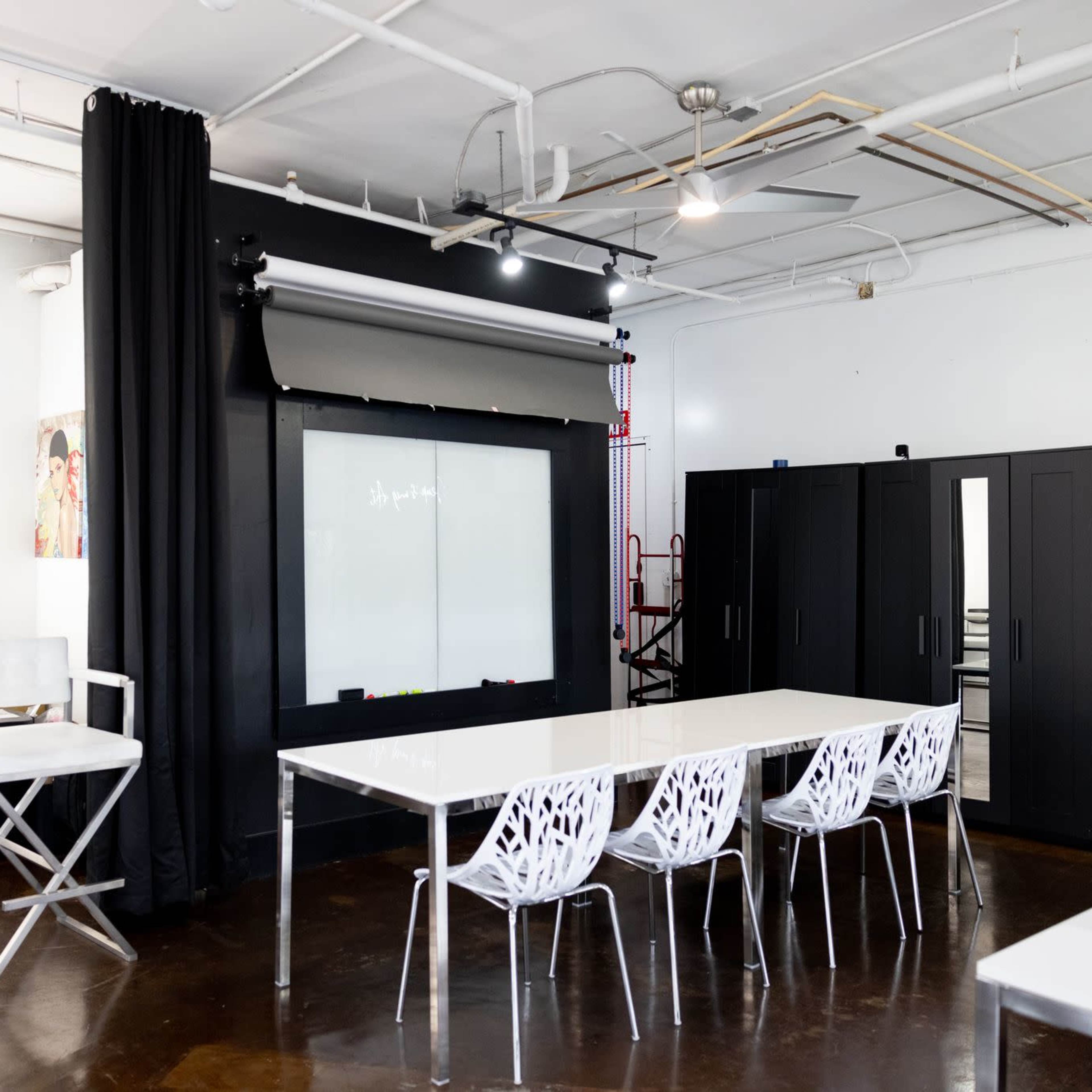 A bright studio space featuring a large table surrounded by white chairs, a backdrop with curtains, and a green wall of plants.