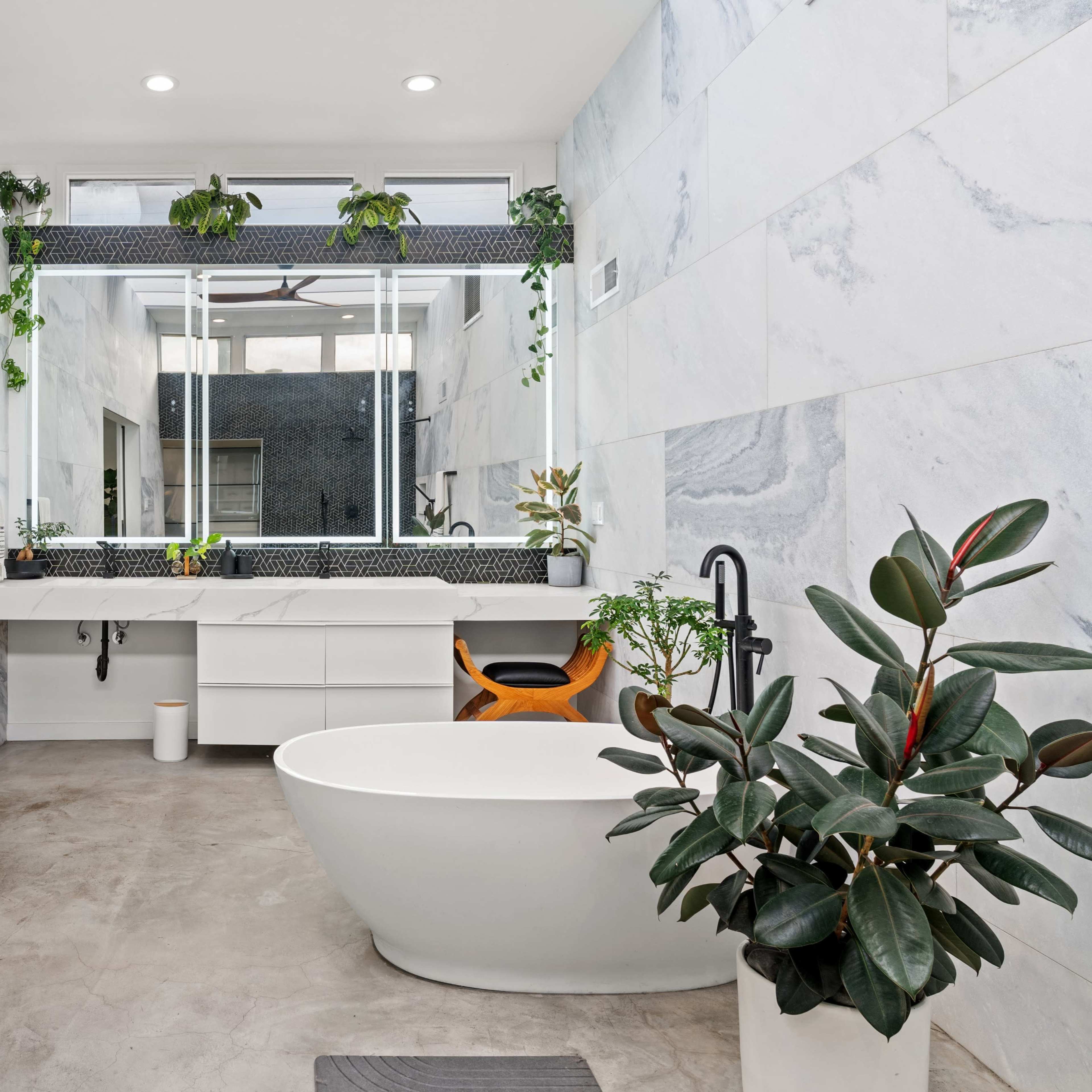 The bathroom features marble walls, a freestanding bathtub, a large mirror, and various indoor plants.