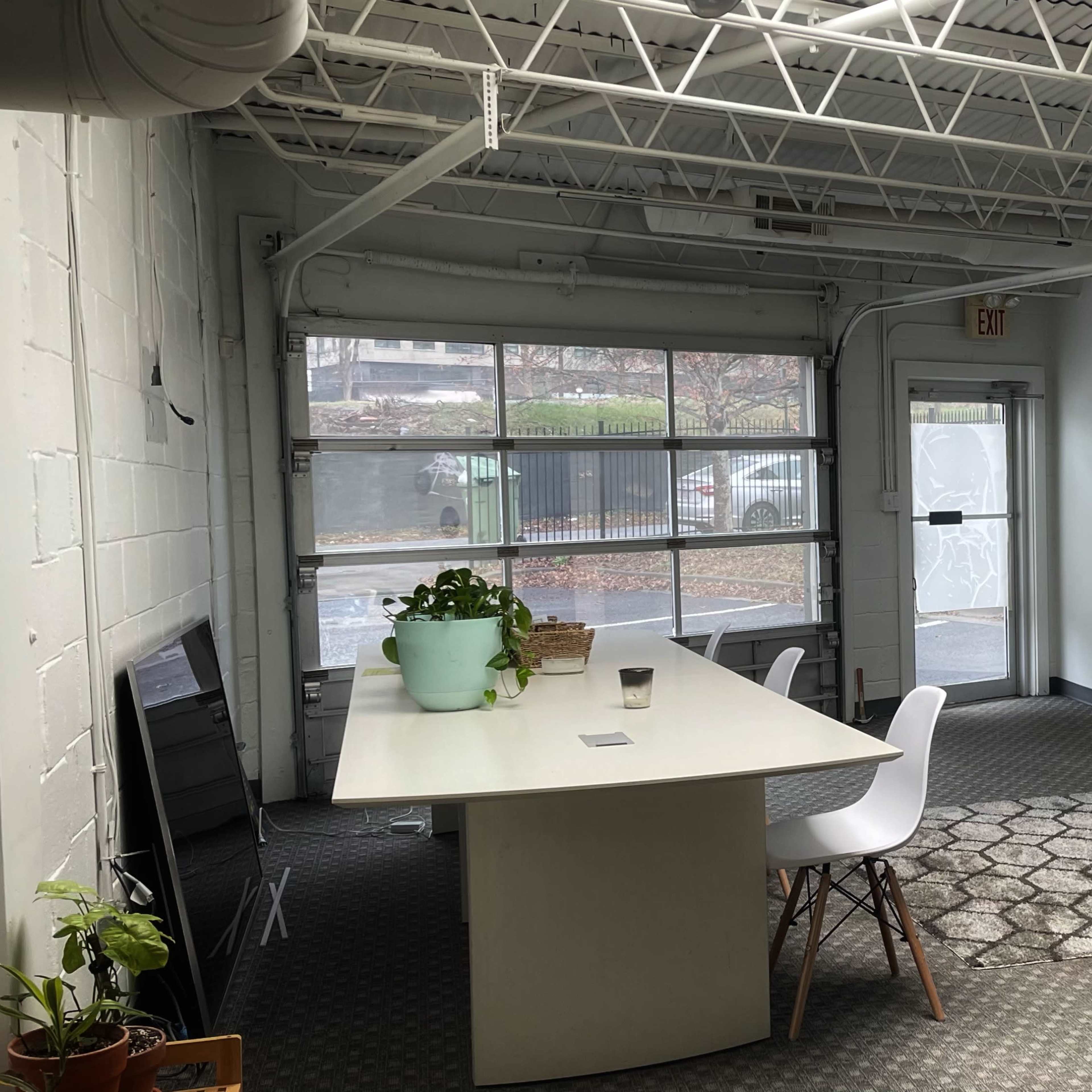 A simple interior space with a large white table, chairs, plants, and windows, along with a door leading outside.