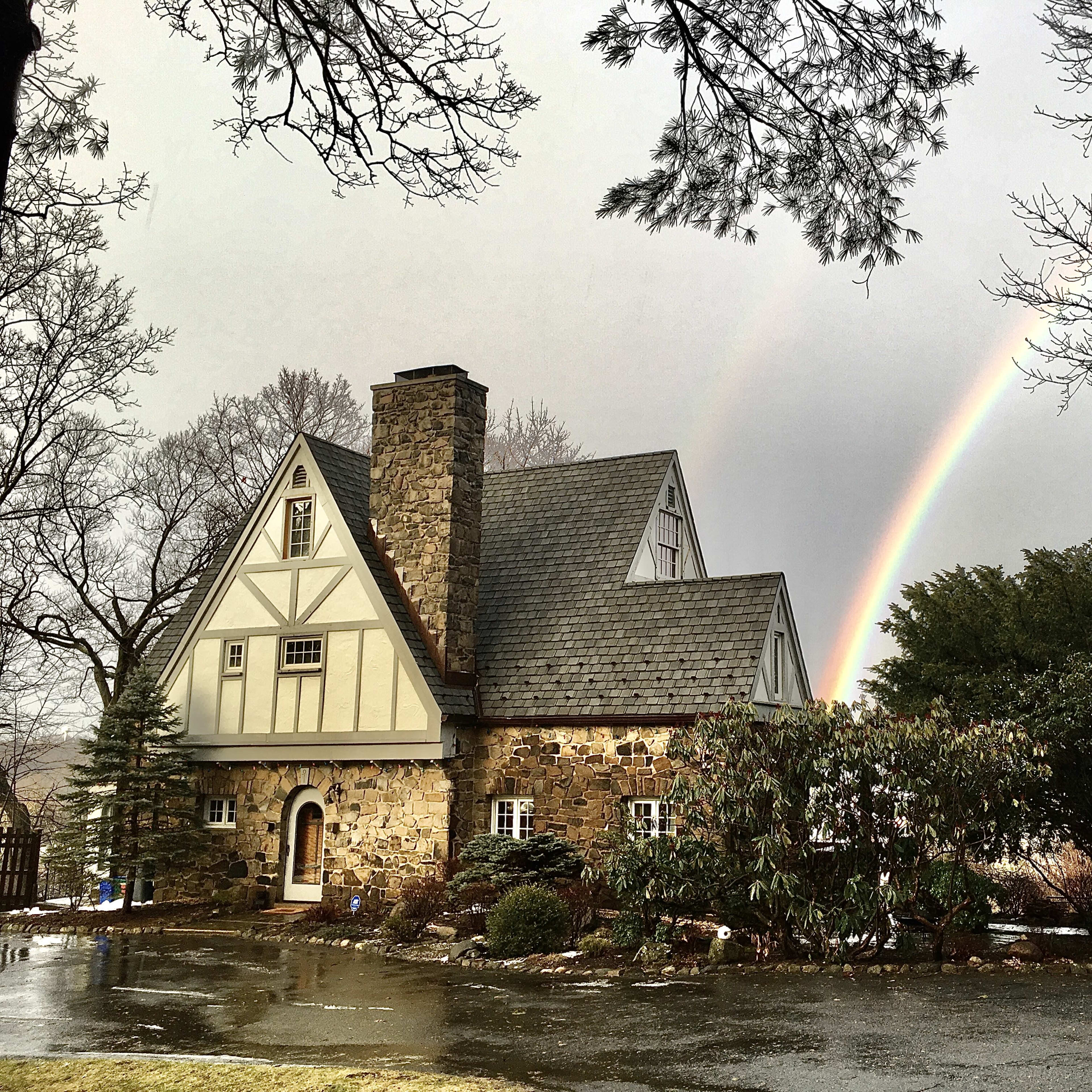 A quaint stone and timber-framed house sits under a gray sky, with a rainbow arcing above it and trees surrounding the property.