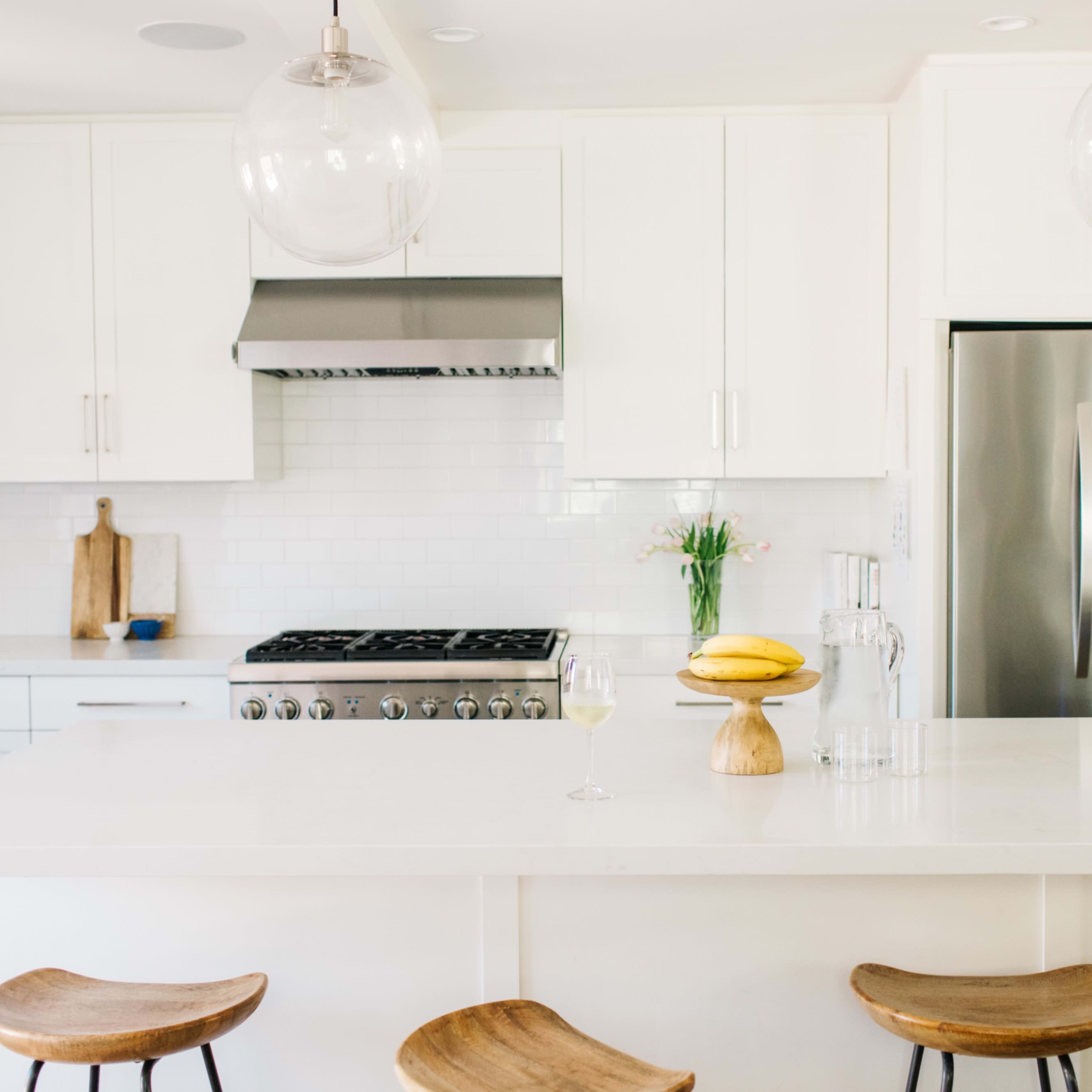 A modern kitchen with white cabinetry, stainless steel appliances, and a central island topped with a wooden display of bananas and a glass.