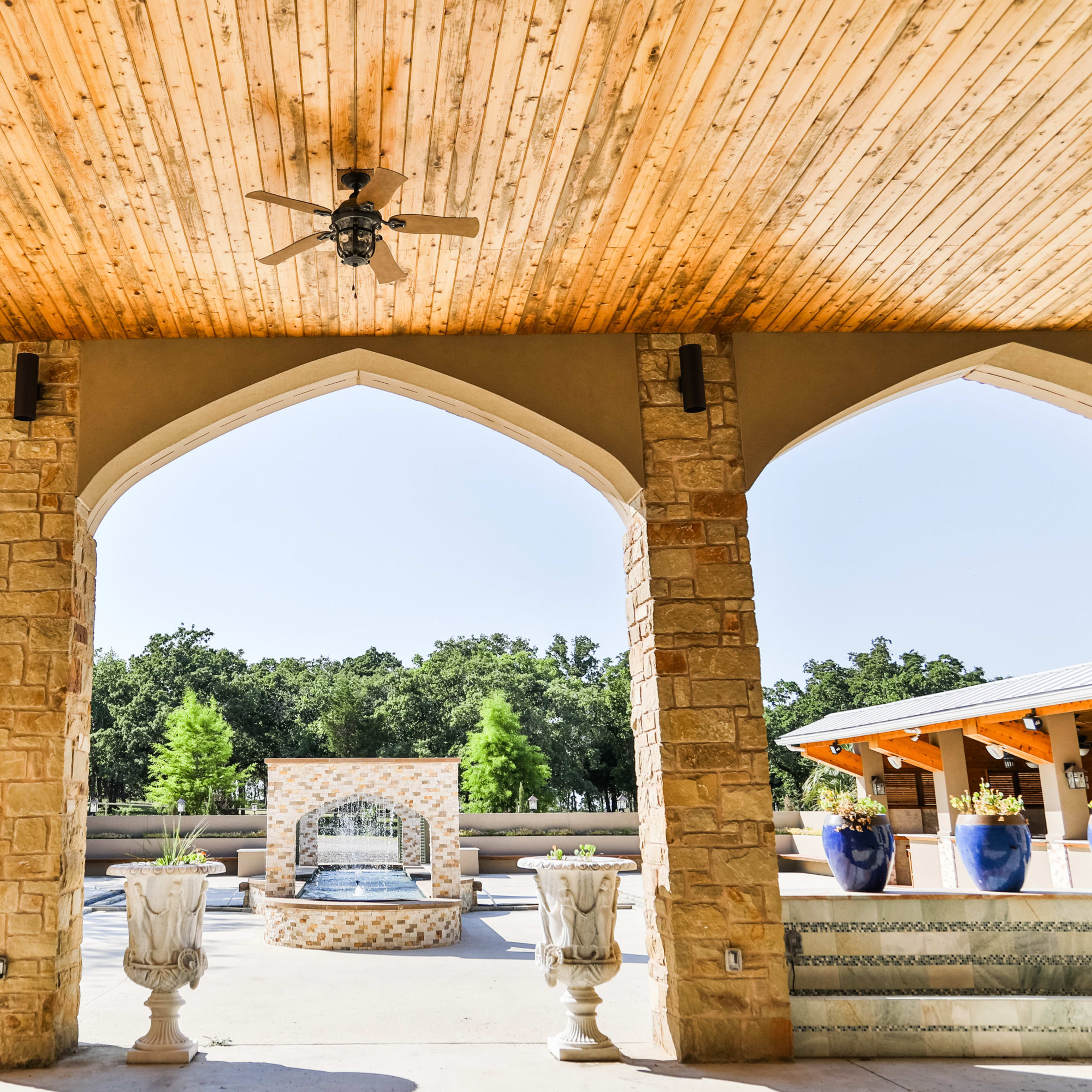 A spacious open pavilion with arched columns, a ceiling fan, and decorative planters, framed by lush green trees in the background.