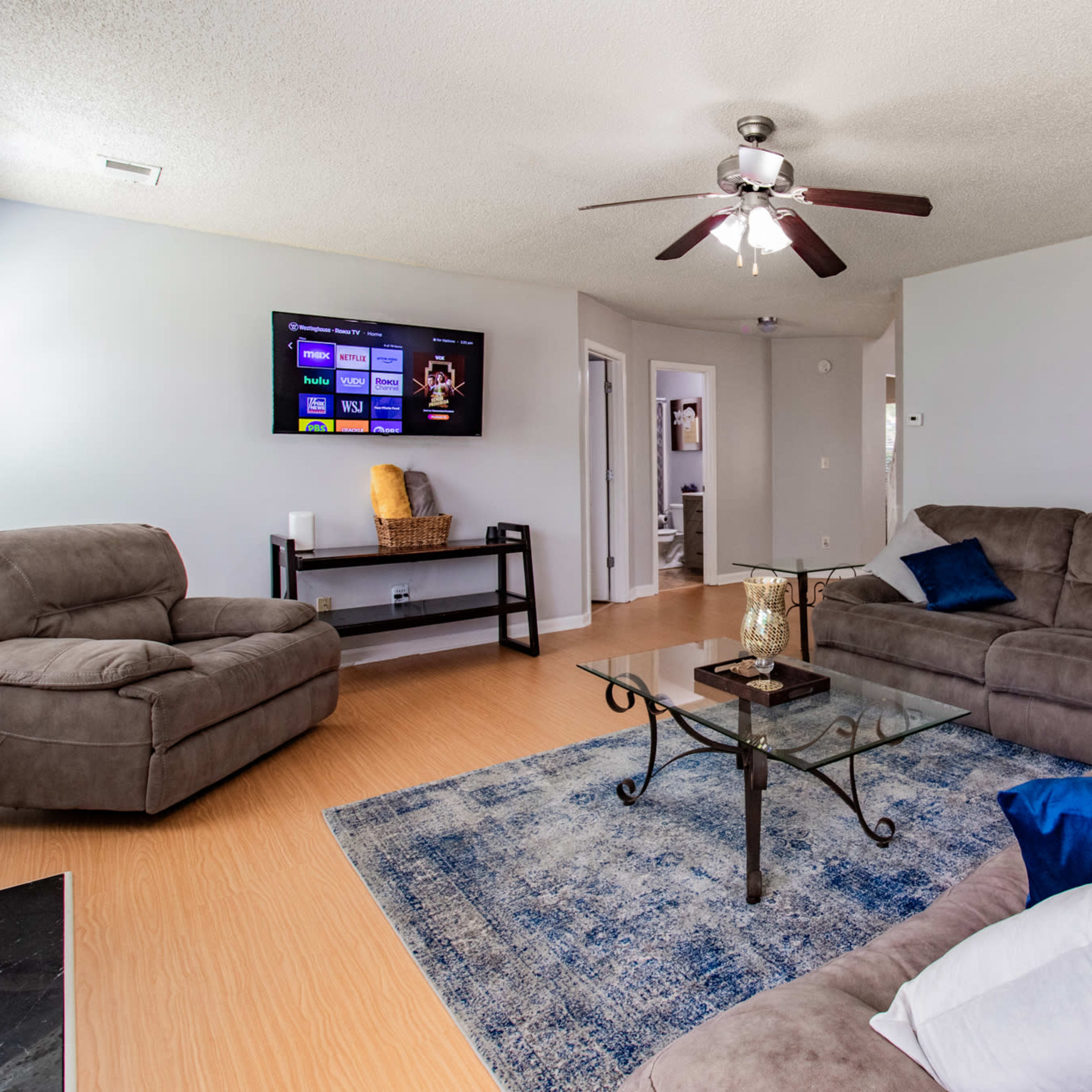 The living room features a sectional sofa, a glass coffee table, and a television mounted on the wall, with a blue area rug on the floor.