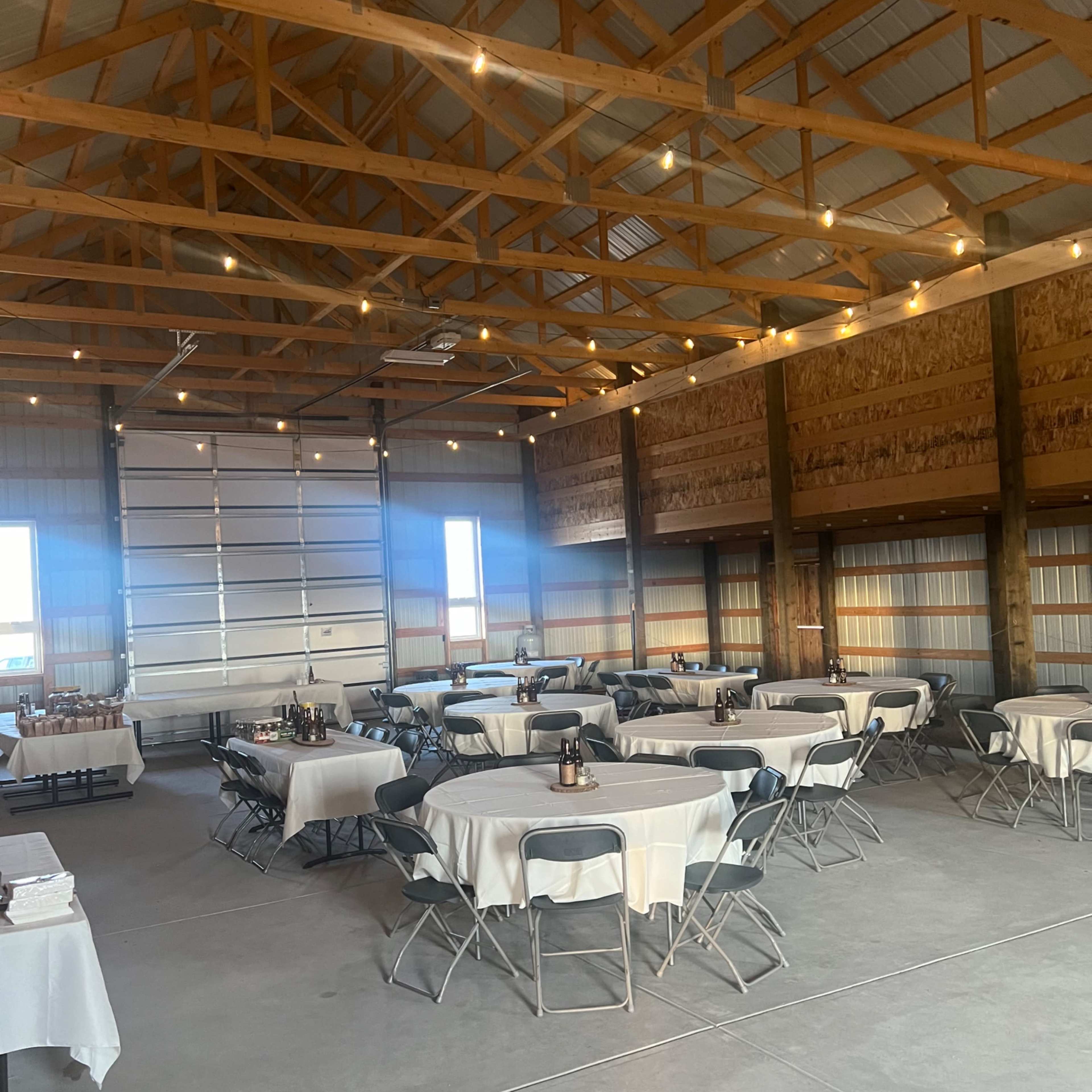 The interior of a spacious barn features several round tables covered with white tablecloths and chairs arranged in an open layout, illuminated by overhead lights.