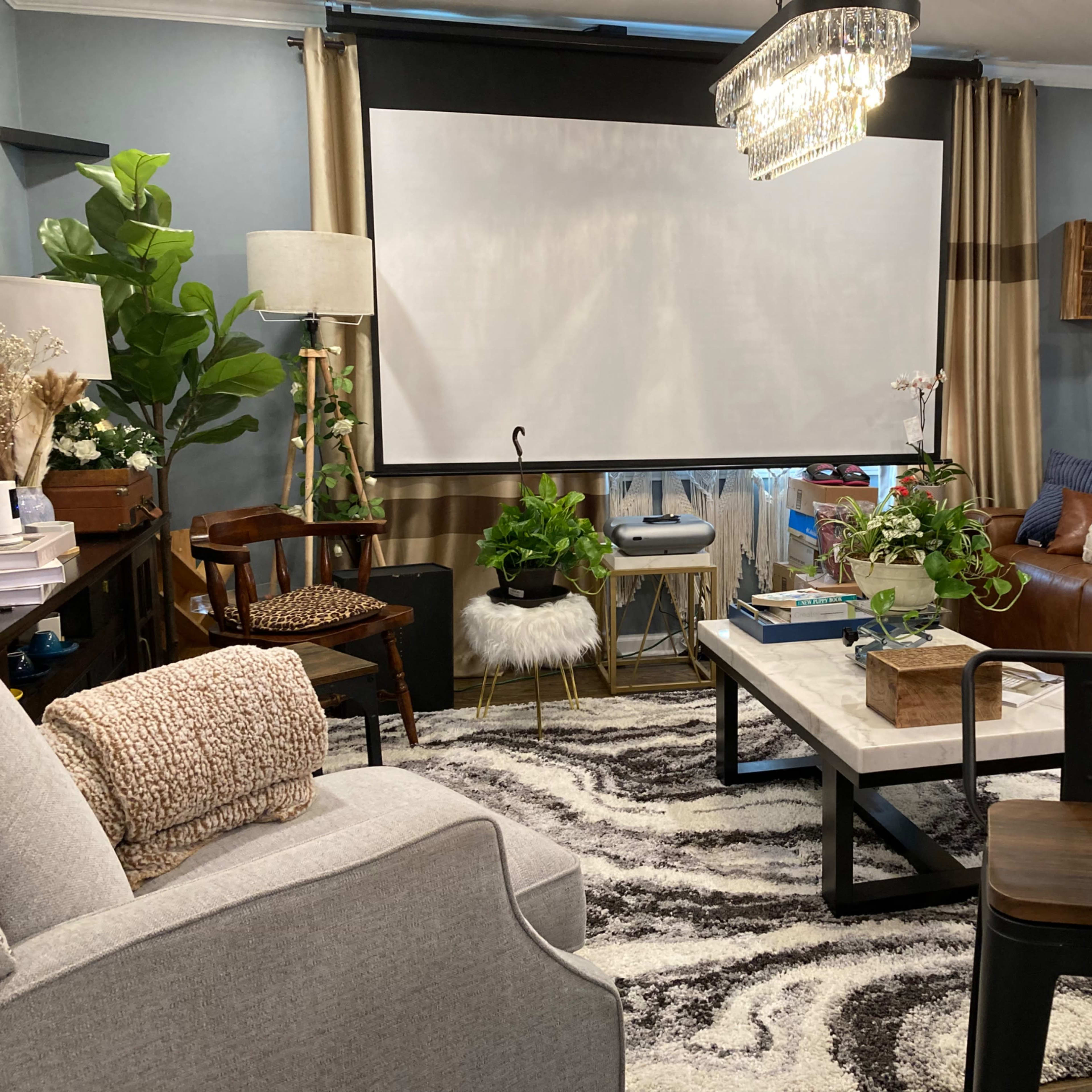 A cozy living room features a large projection screen, comfortable seating, decorative plants, and a patterned area rug.