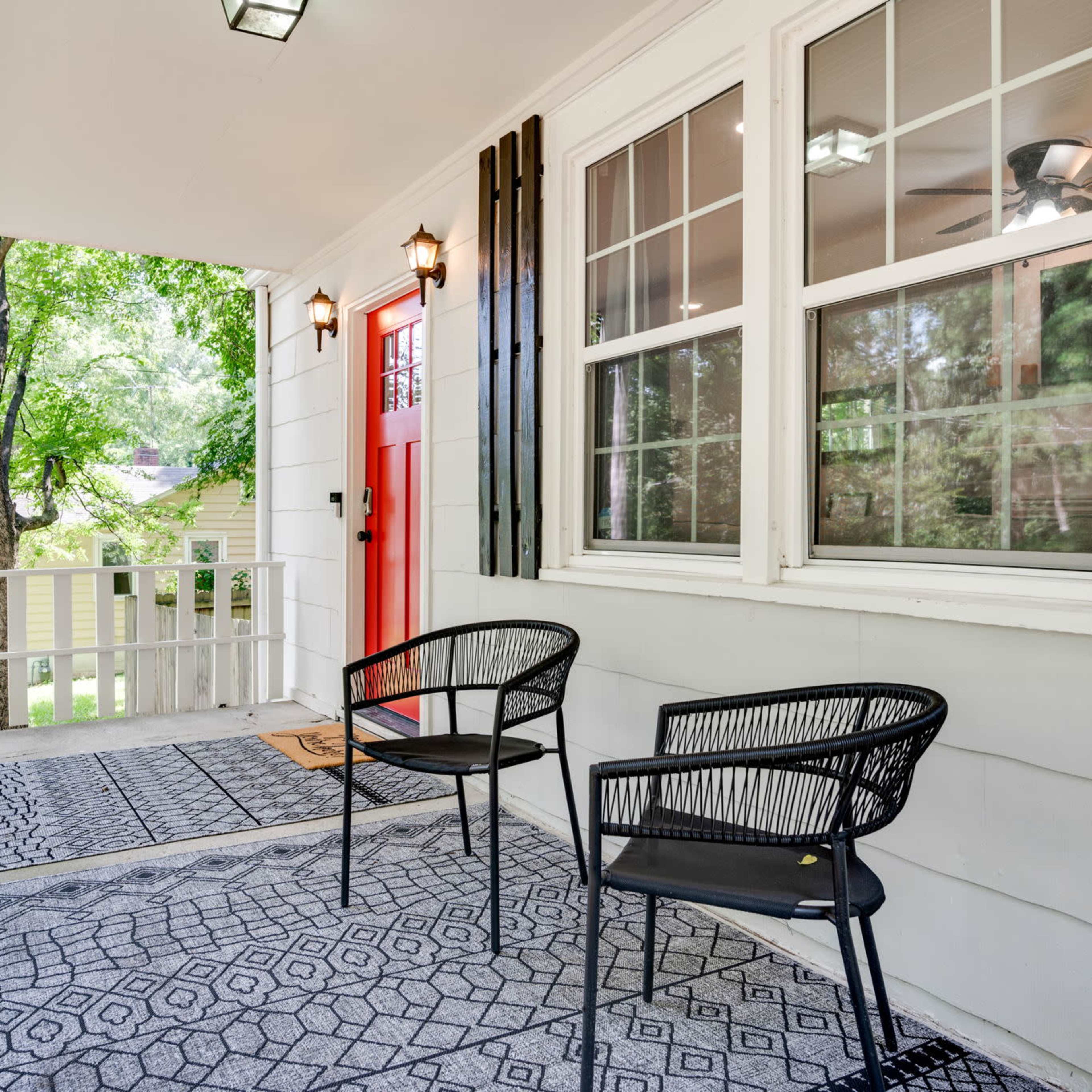 A porch with two black chairs, a red front door, and patterned rugs under a covered area surrounded by greenery.
