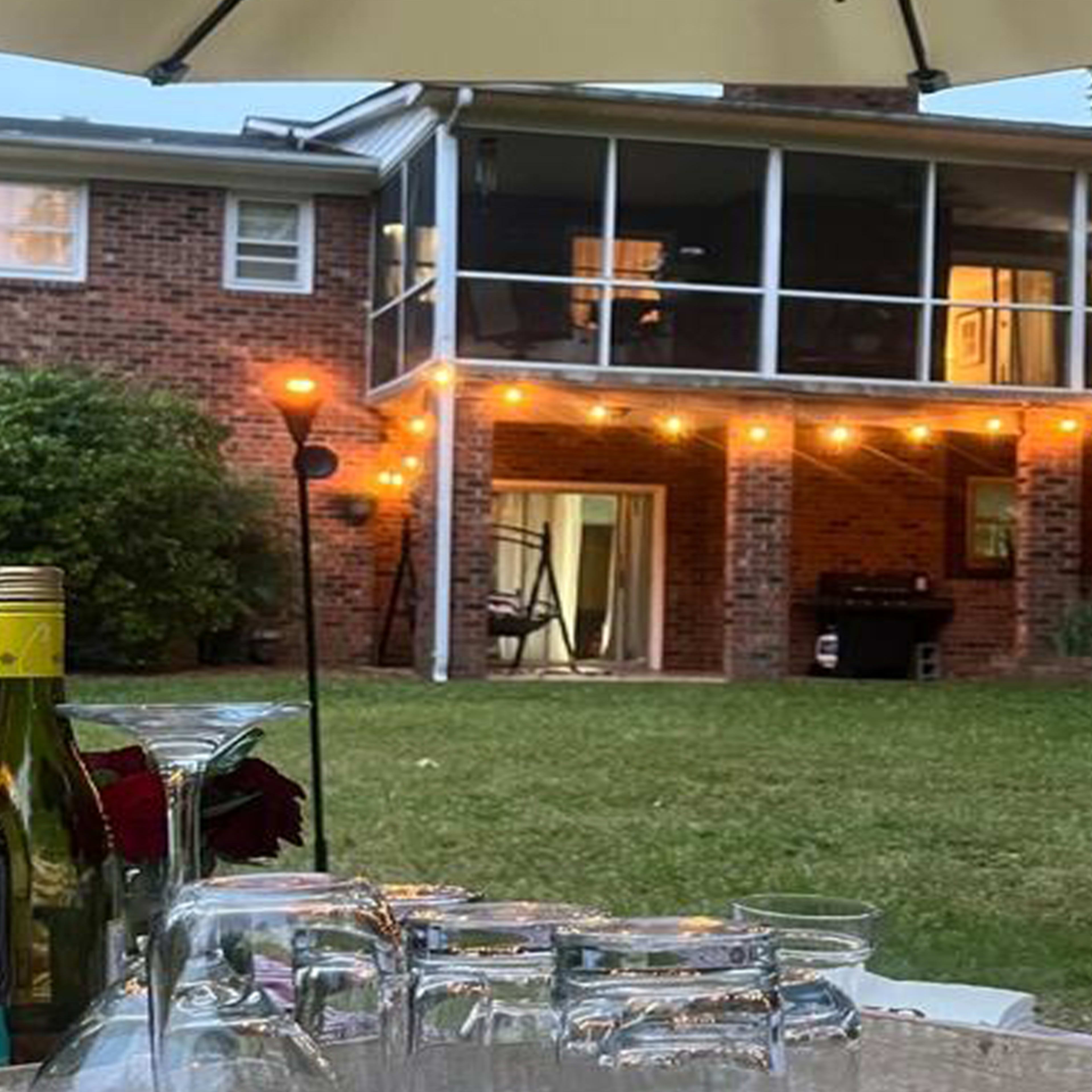 A backyard with a table set with wine bottles and glasses, while a two-story brick house with a screened porch is illuminated by string lights.