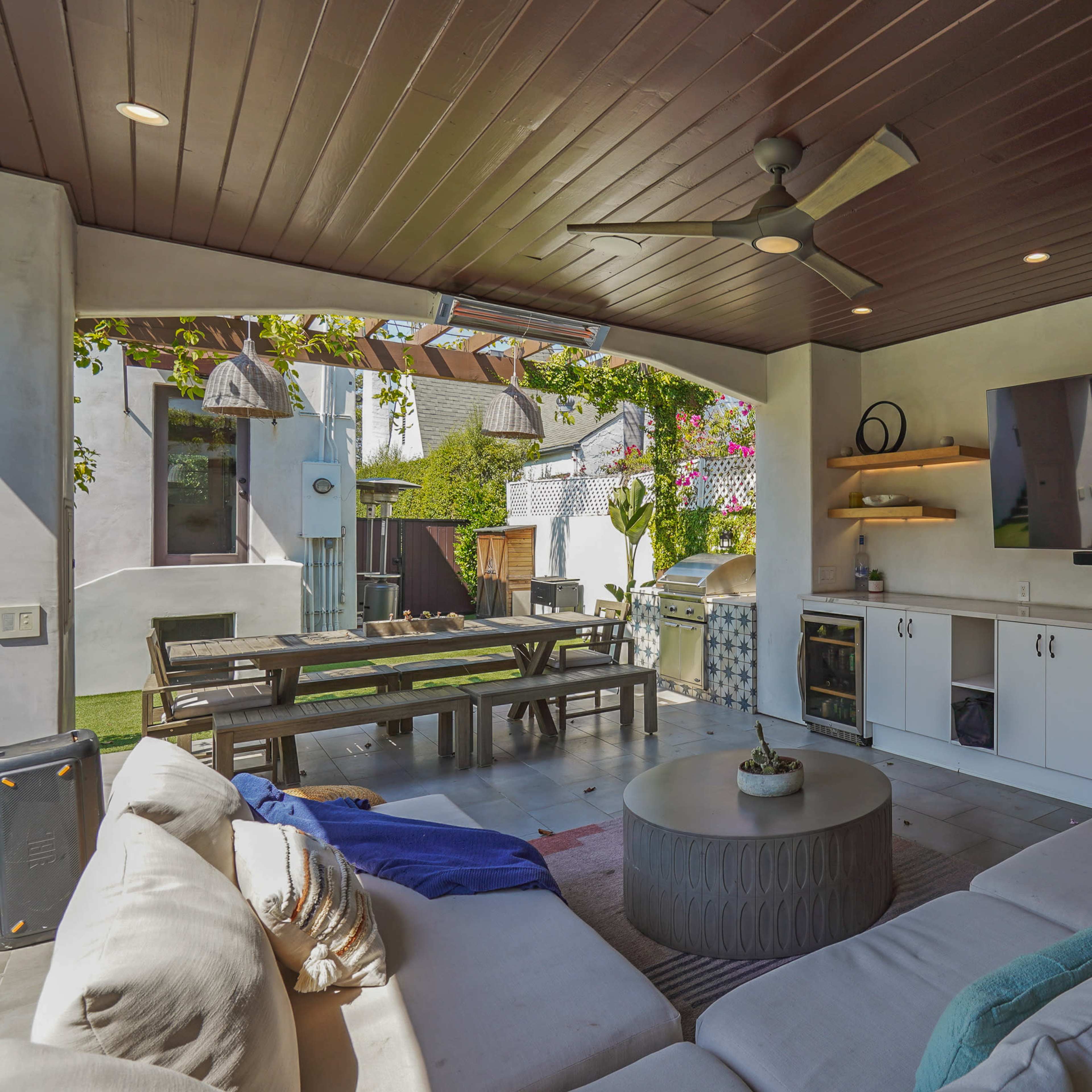 A modern outdoor living space featuring a sofa, a mounted television, and a dining area surrounded by greenery.