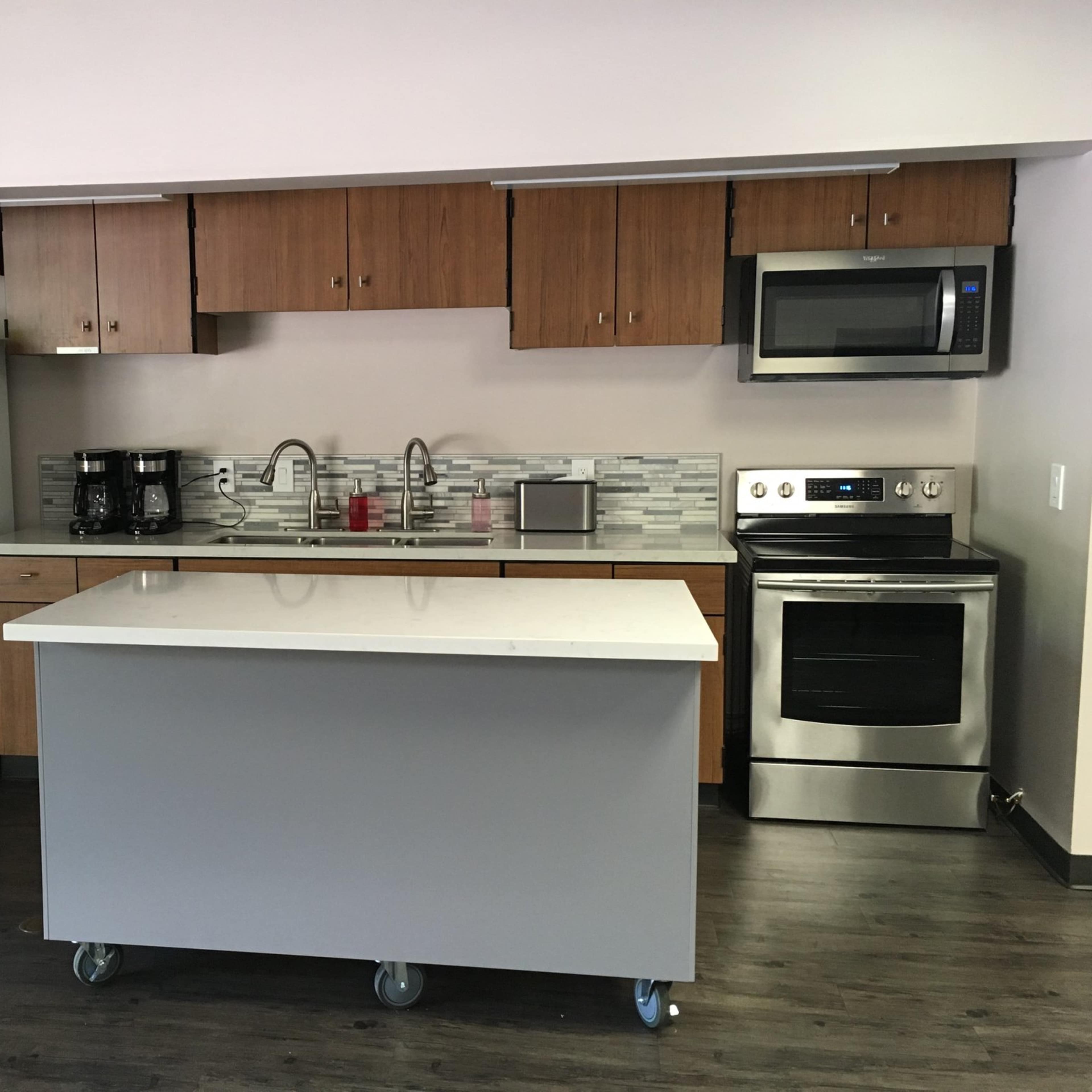 A modern kitchen with wooden cabinets, stainless steel appliances, a white countertop island on wheels, and a tiled backsplash.