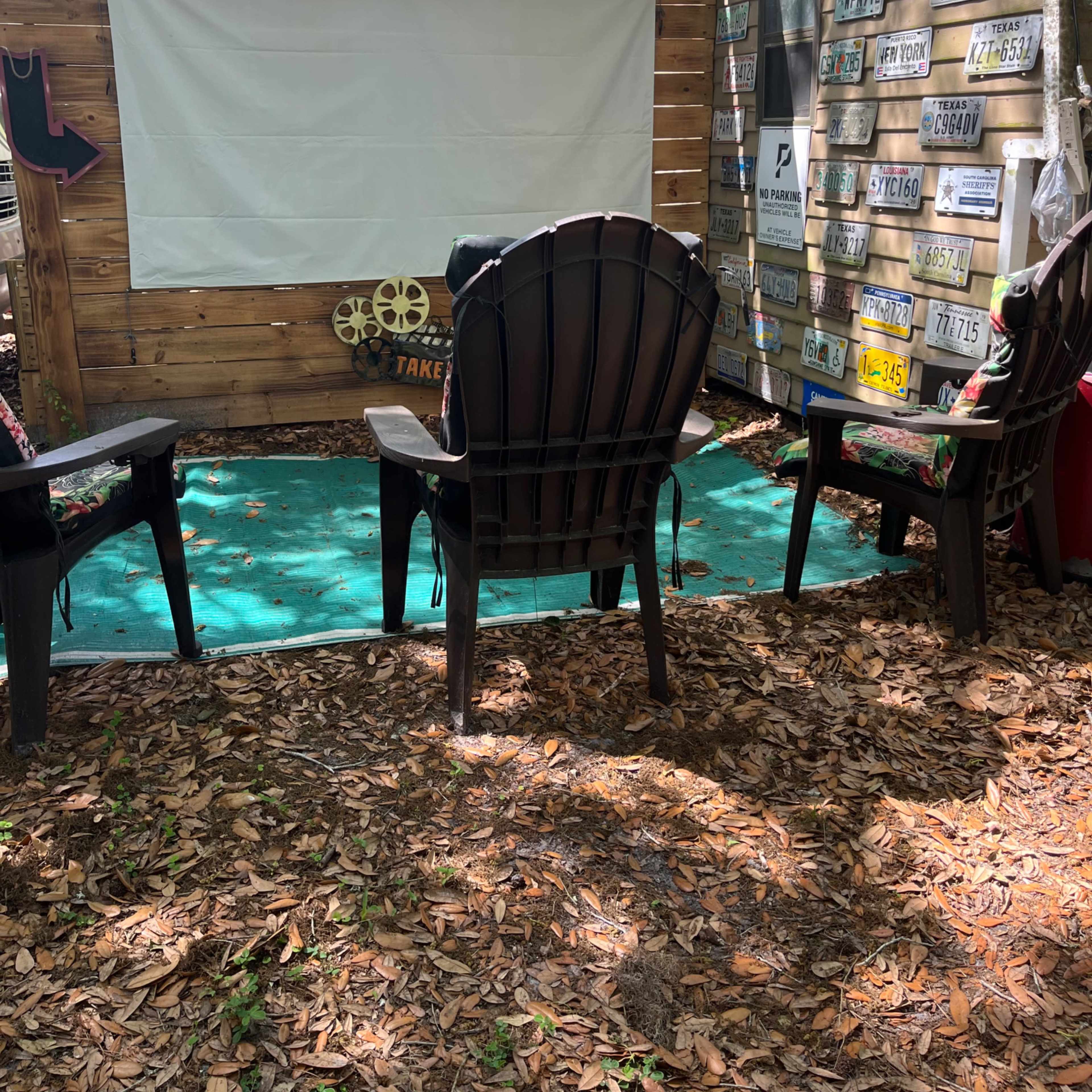 The scene features an outdoor sitting area with black adirondack chairs facing a white canvas screen, surrounded by a wooden backdrop and a wall adorned with various license plates.