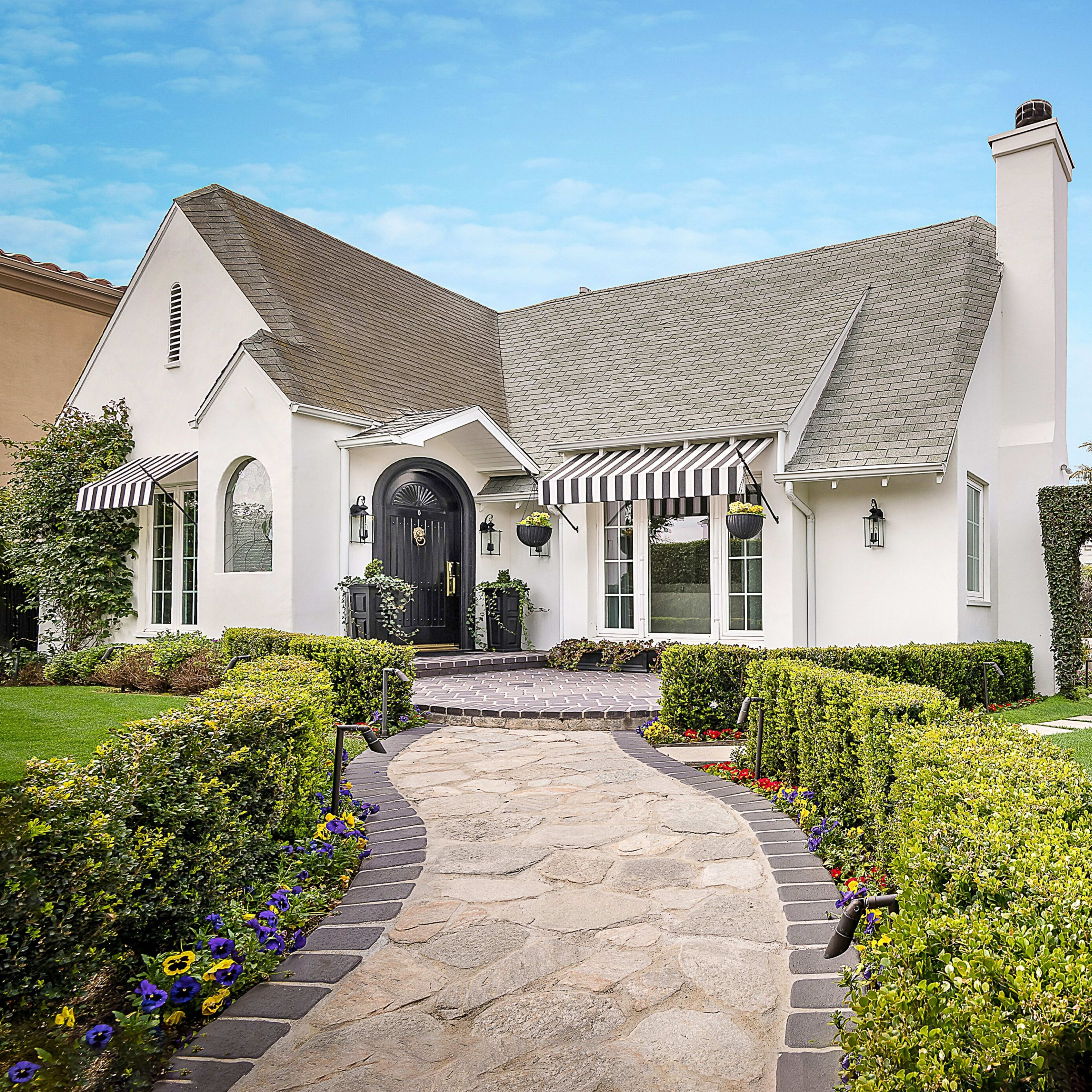 A white house with a grey roof and black door is surrounded by neatly trimmed hedges and a stone pathway leading to the entrance, with lemon trees visible on the right.