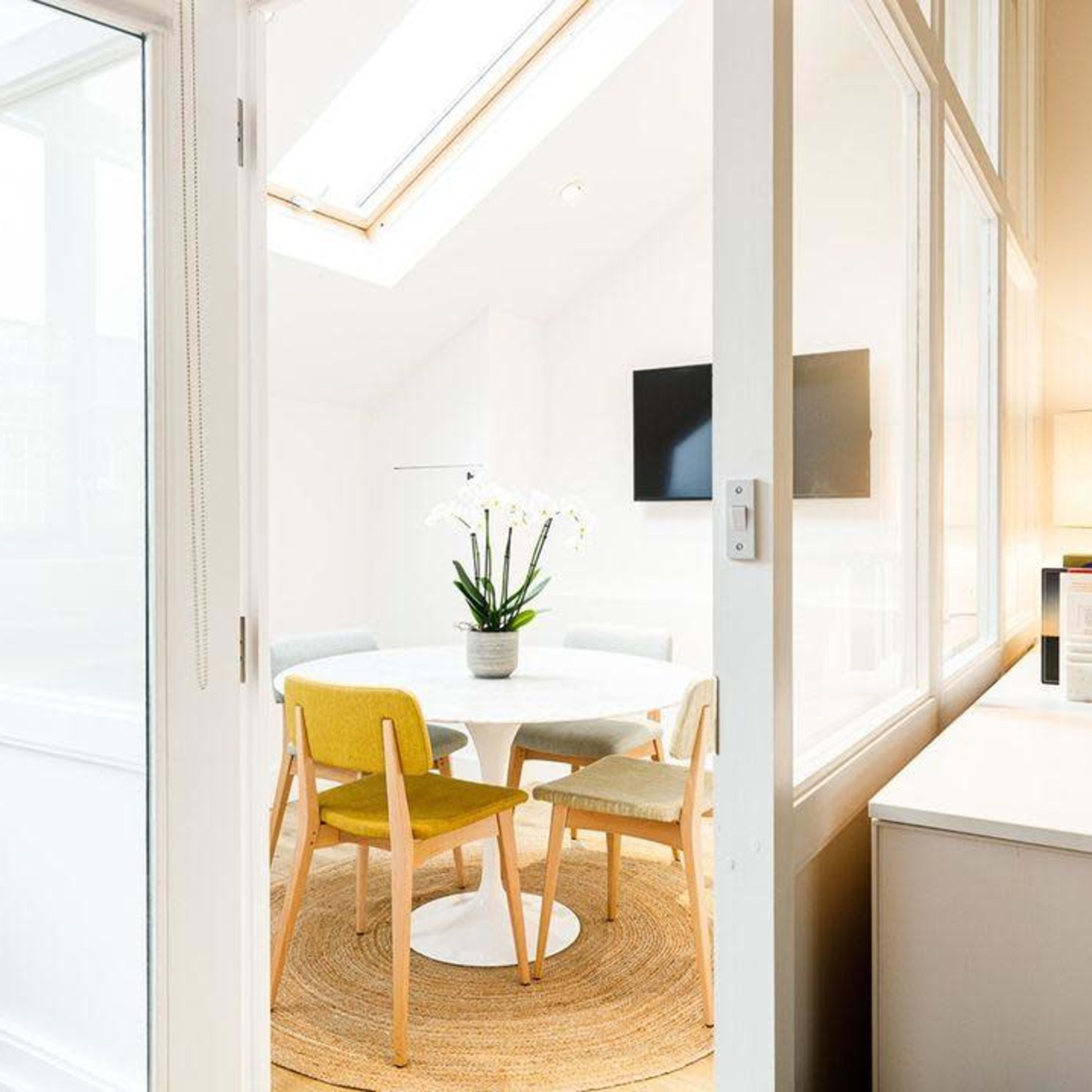 A bright room features a round table surrounded by chairs, with a plant centerpiece and a lamp on a nearby side table.