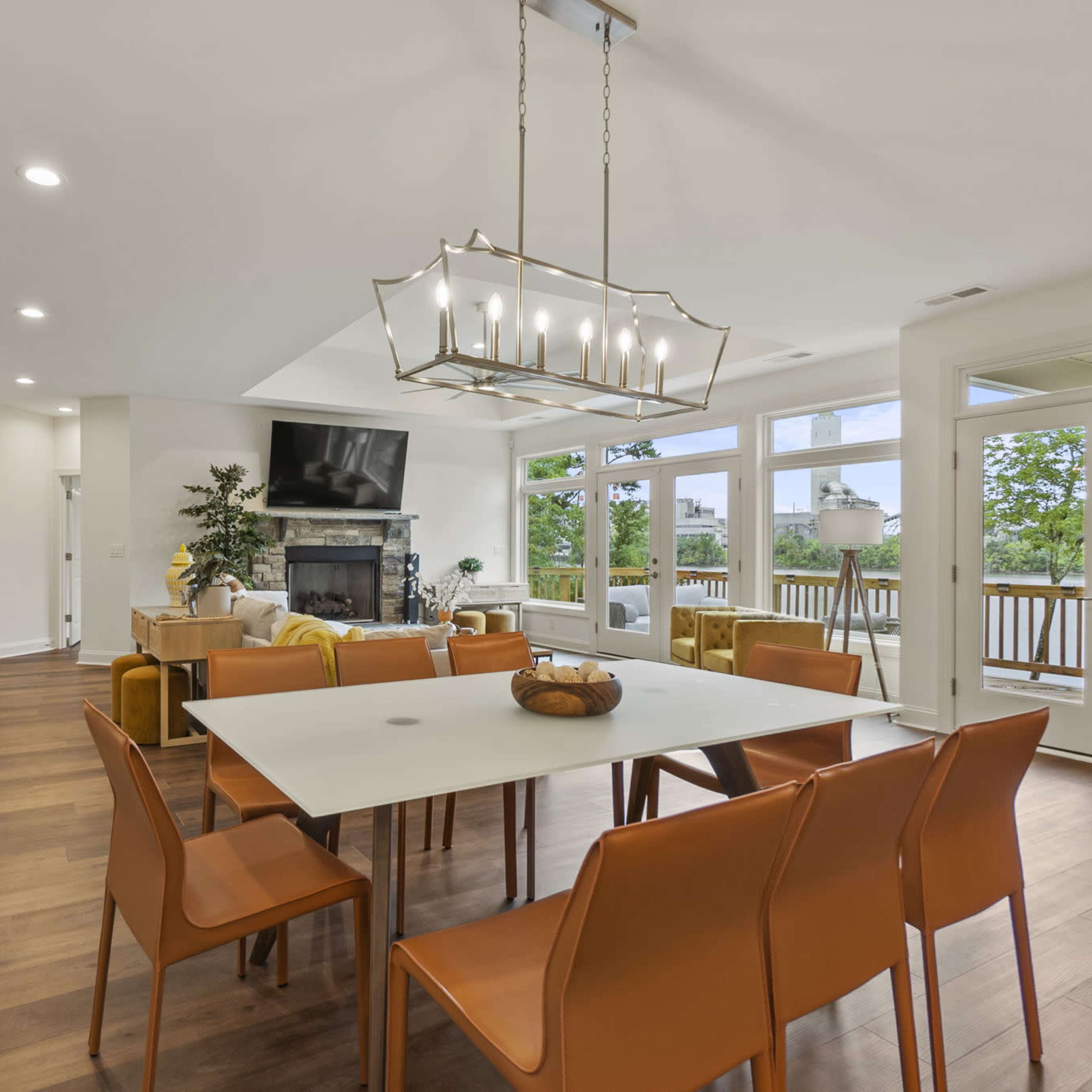 A modern dining area features a white table surrounded by orange chairs, adjacent to a living space with a large window and a fireplace.