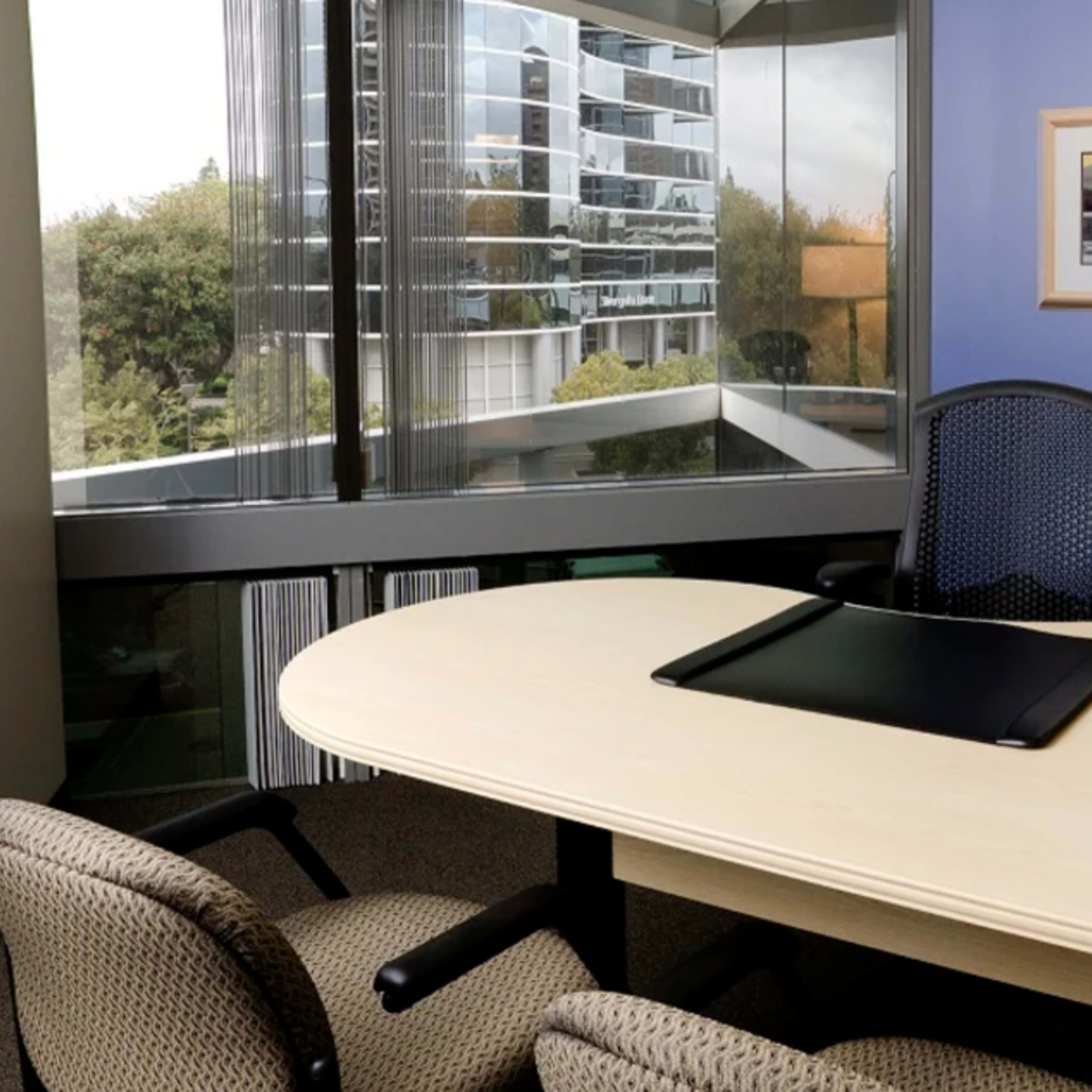 A modern office space featuring a large desk, an office chair, a lamp, and a window view of a multi-story building and greenery outside.