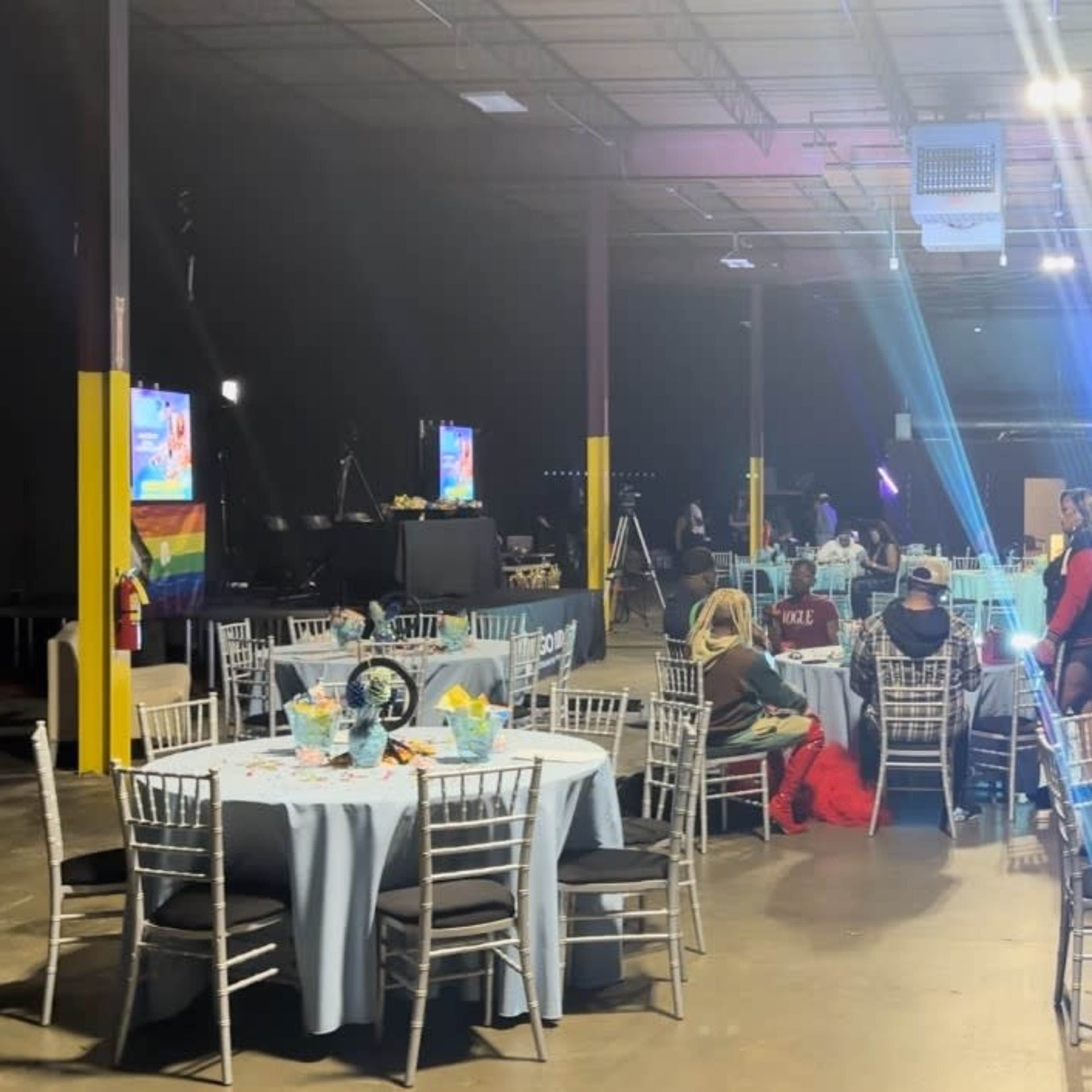 An event space with multiple round tables set for dining, decorated with colorful centerpieces, and illuminated by overhead lights.