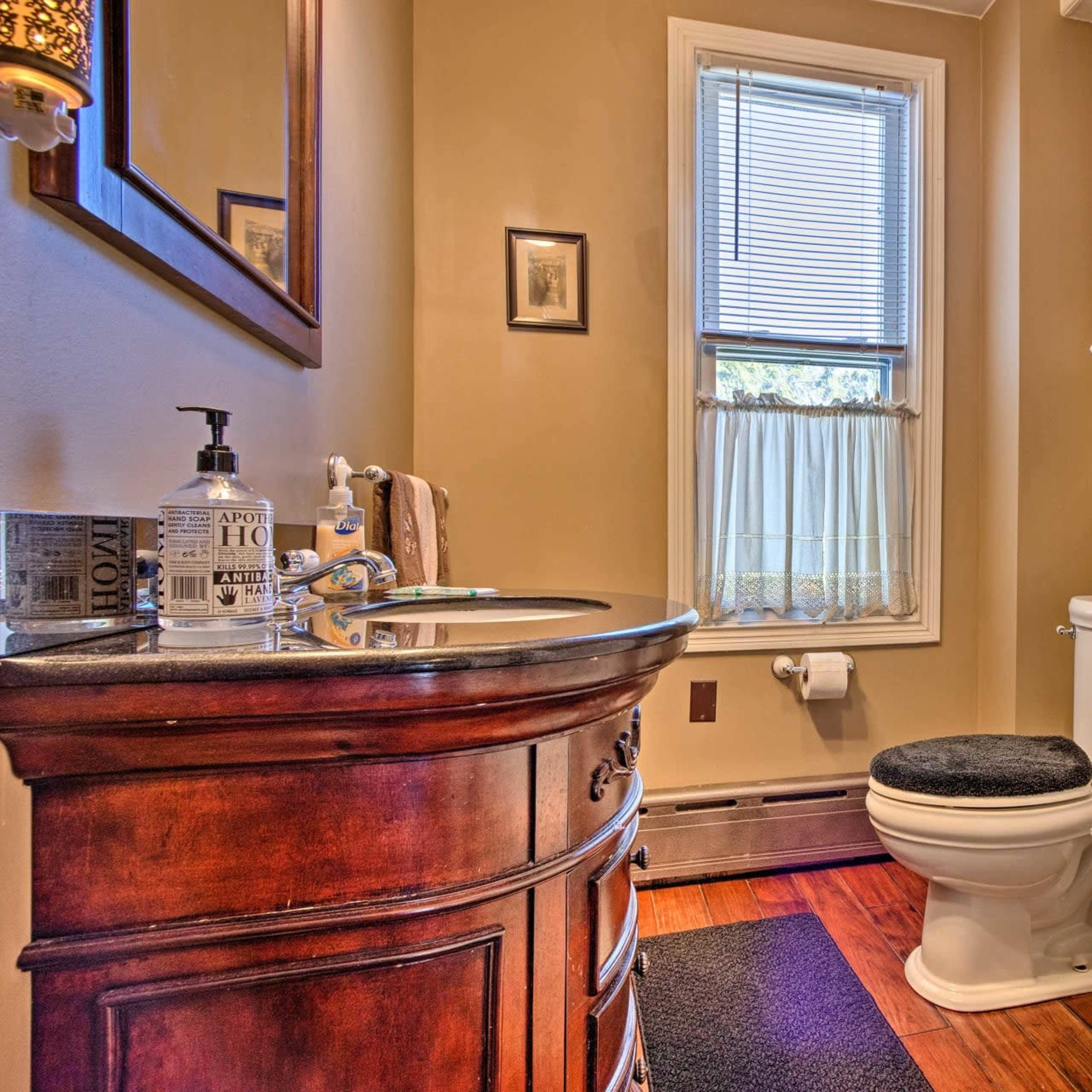 A small bathroom with a wooden vanity, a toilet, a bathtub, and a window with sheer curtains.