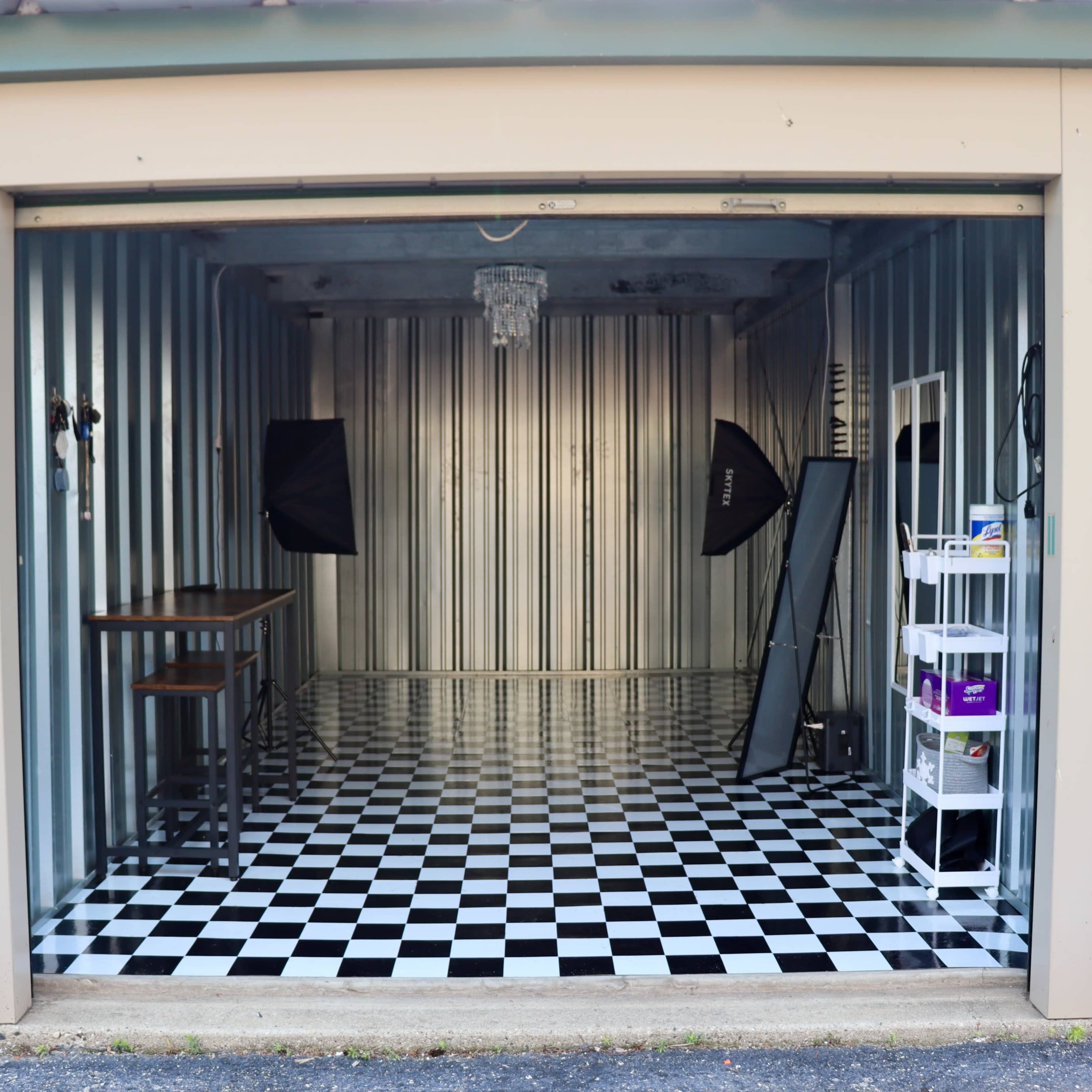 An empty storage unit with a black and white checkered floor, reflective walls, and a small table set up with lighting equipment.