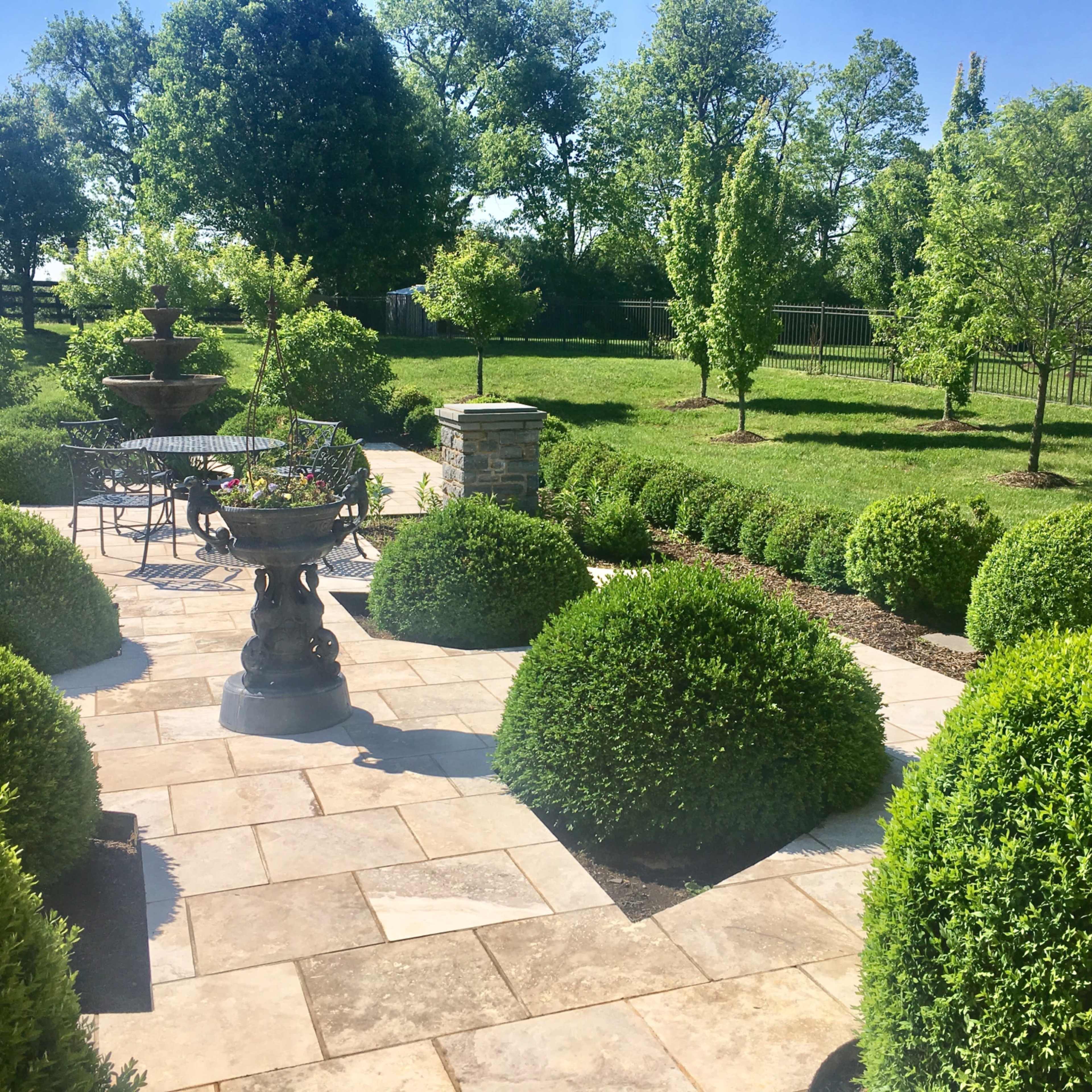 A well-maintained patio features circular hedges, a stone fountain, and a metal table with chairs, surrounded by a green lawn and trees.