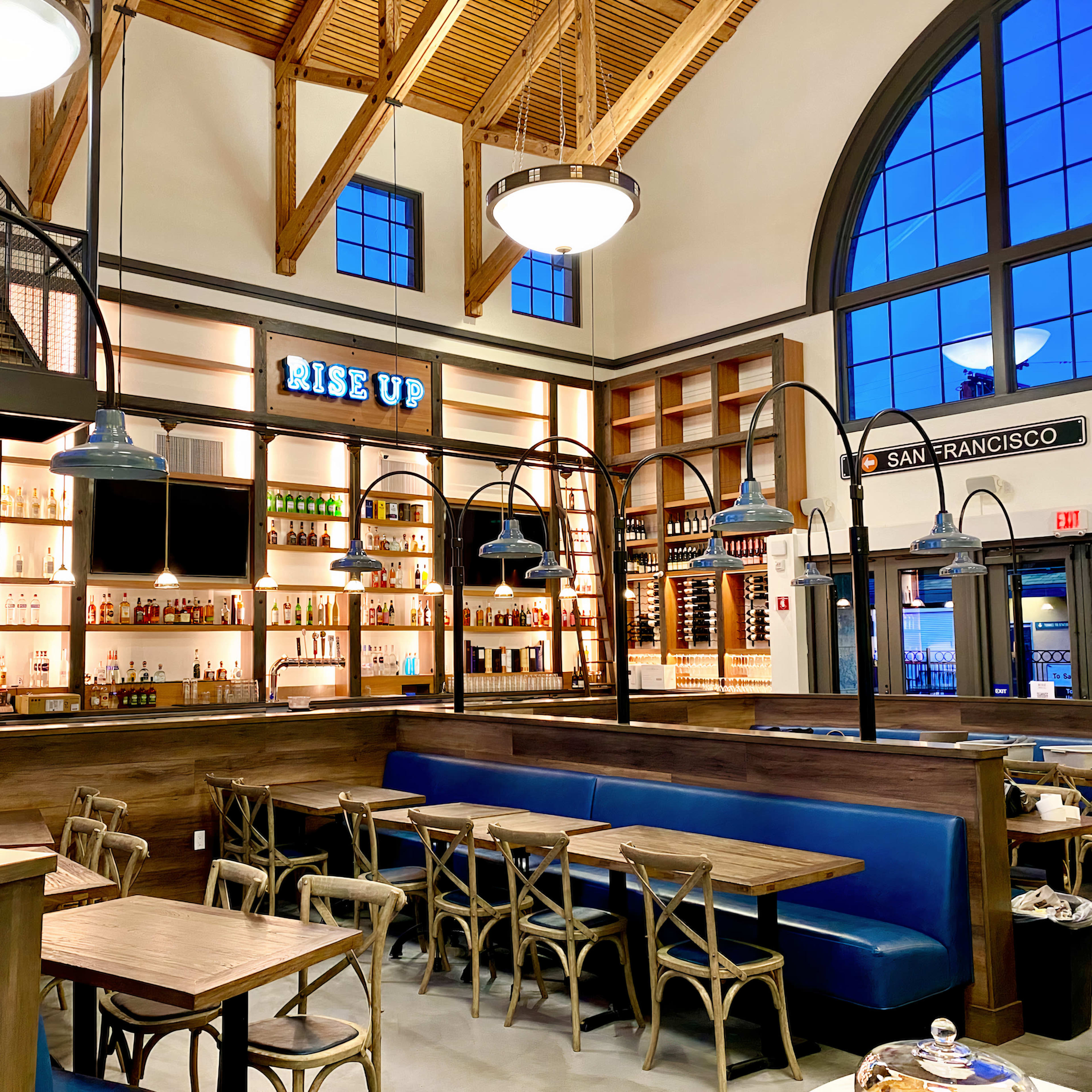 A modern restaurant interior with wooden furnishings, blue booth seating, and a bar displaying various bottles, all under a high ceiling with large windows.