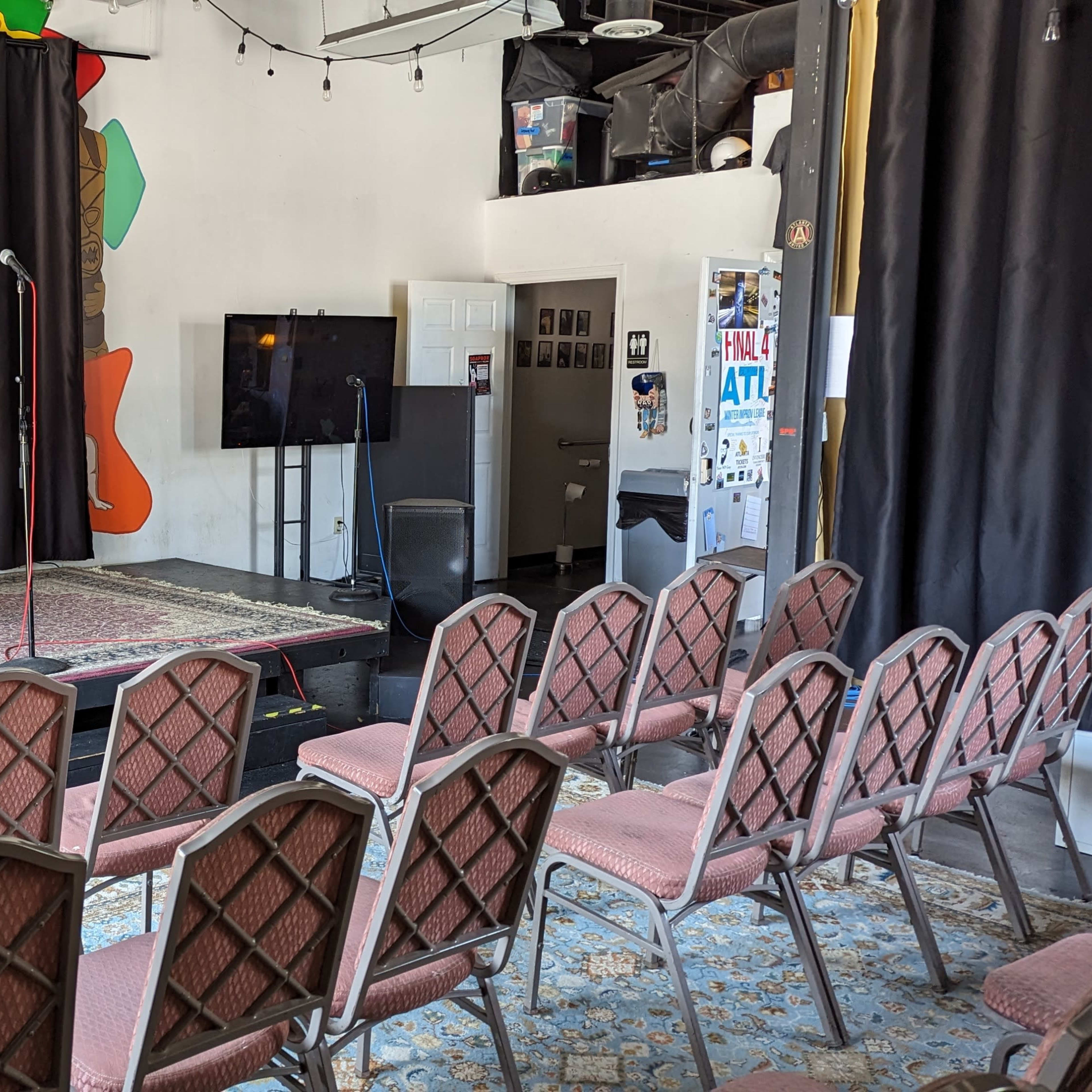 An empty stage with a microphone and a folding chair facing an audience area filled with rows of seats.