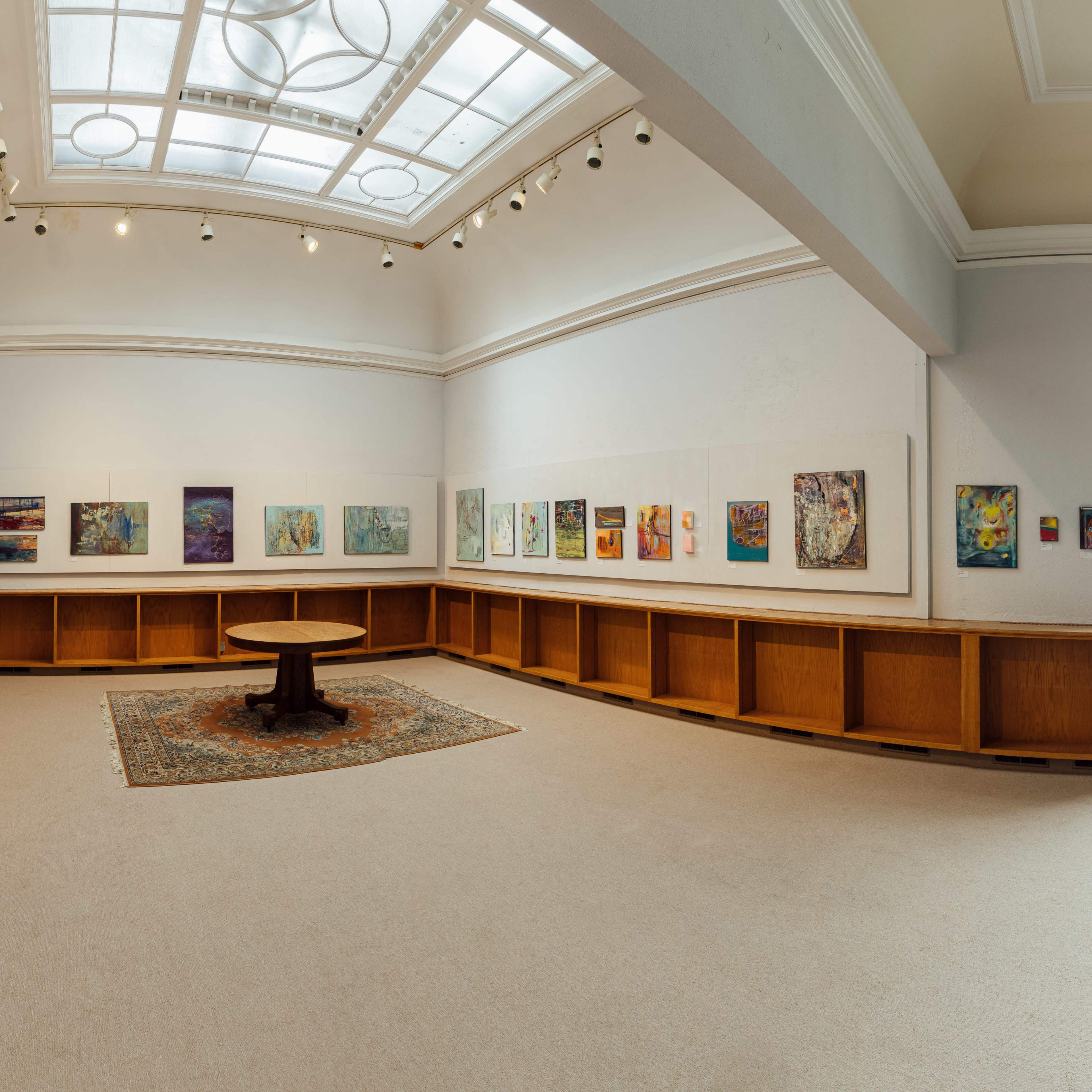 An art gallery featuring colorful paintings displayed on the walls, with a central round table on a patterned rug.