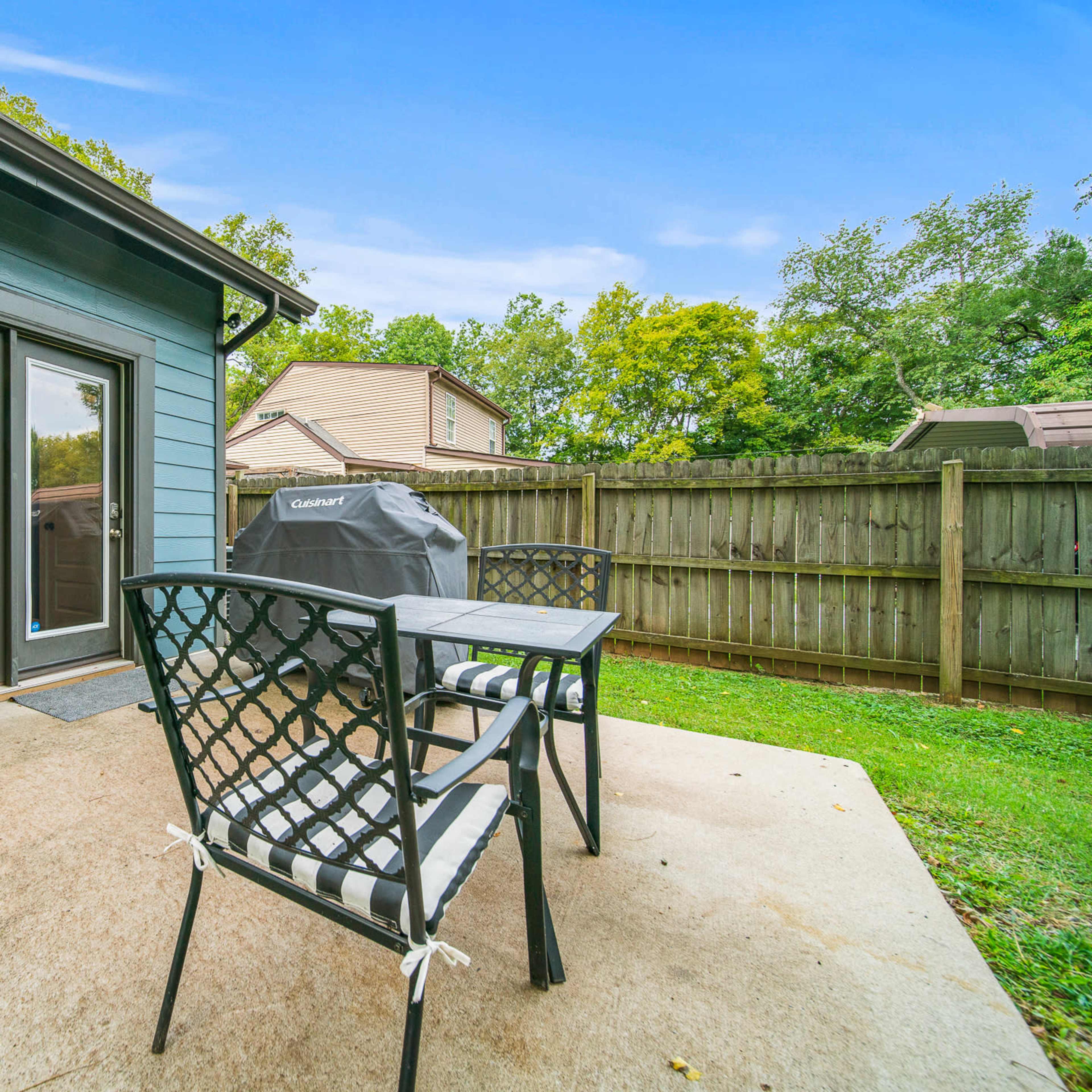 A backyard patio with two black chairs and a grilling area, surrounded by a wooden fence and greenery.