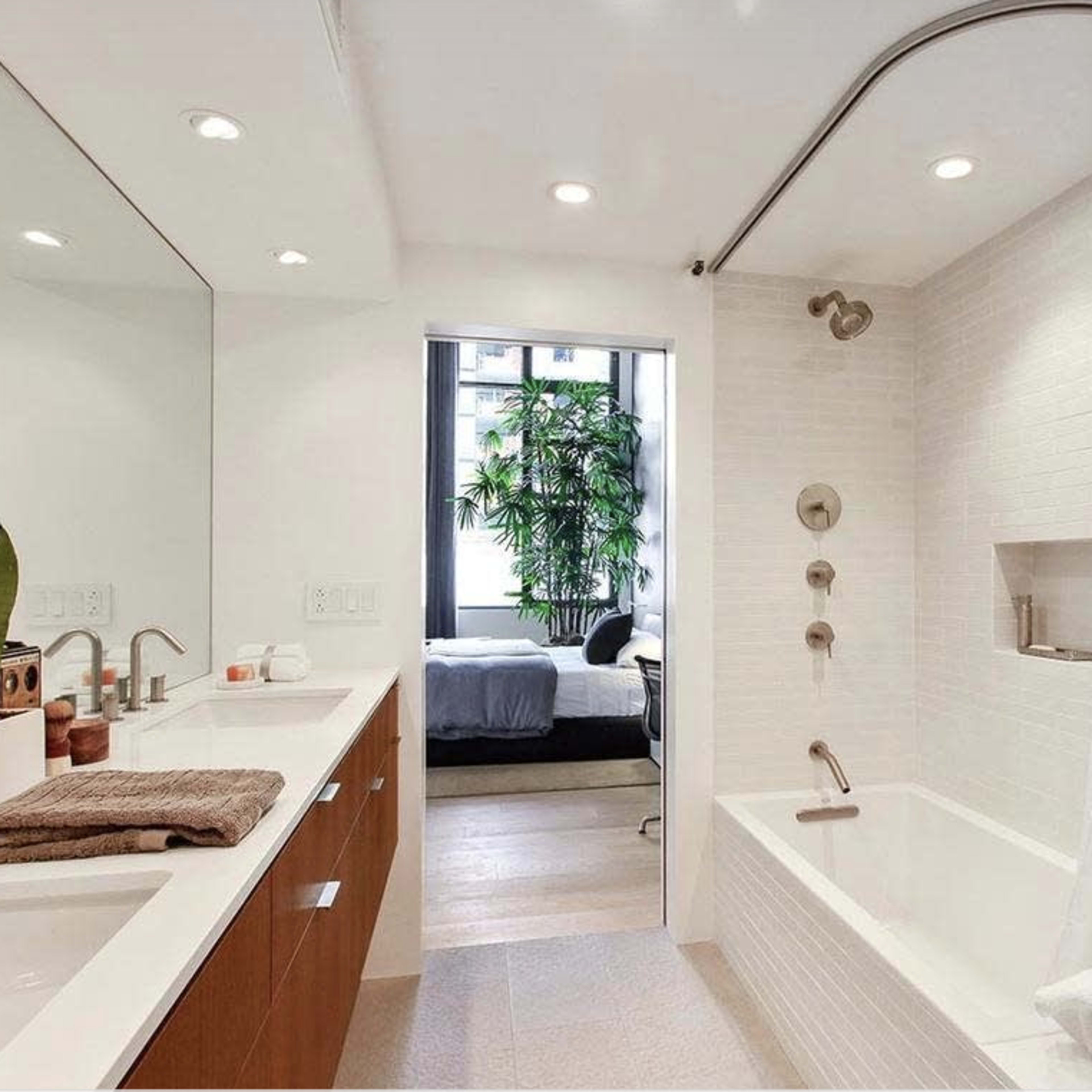 A modern bathroom with a bathtub, a shower curtain, a vanity with two sinks, and a large mirror, with greenery visible through a window.