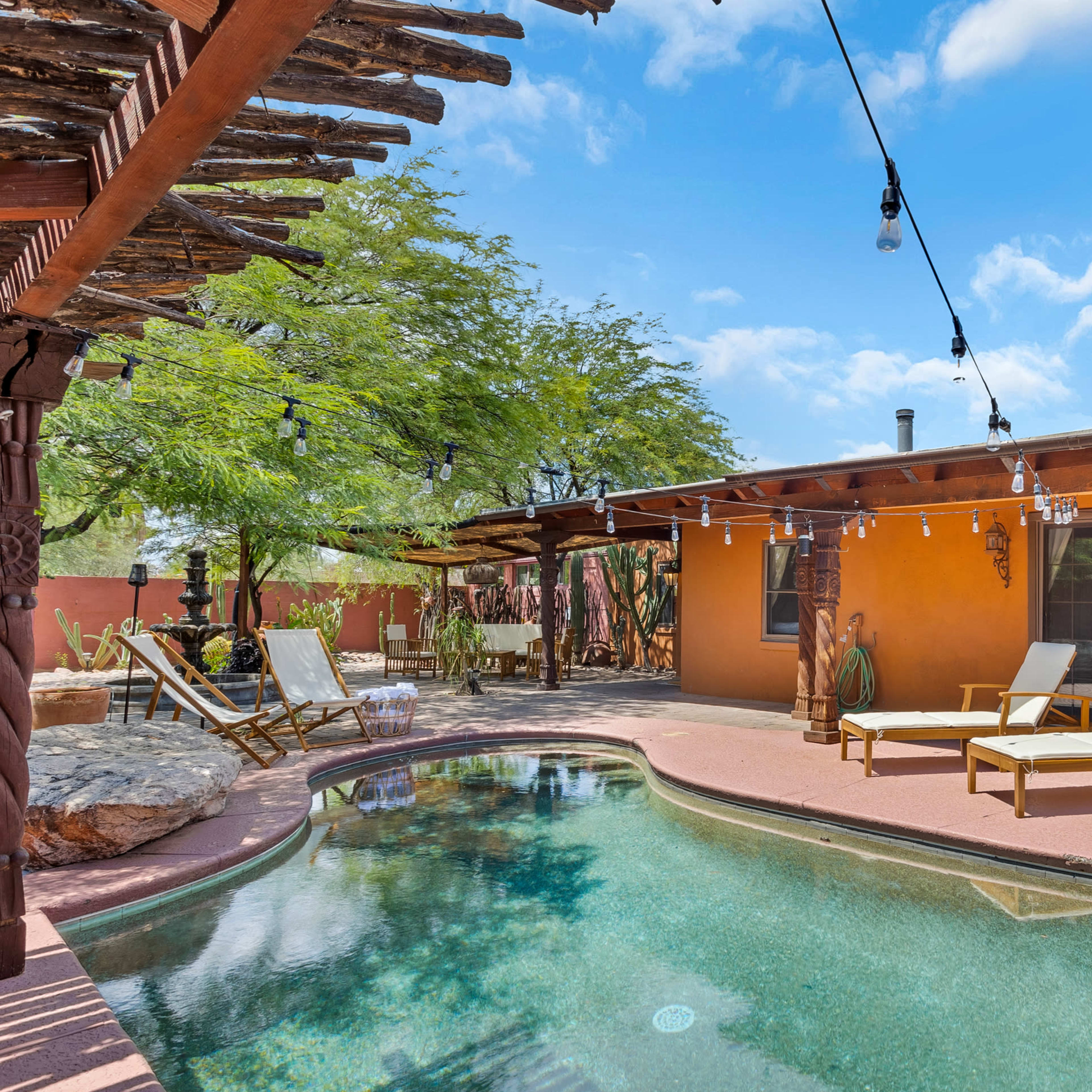 A backyard pool area with two lounge chairs beside a curved swimming pool, surrounded by desert-style landscaping and festive string lights overhead.
