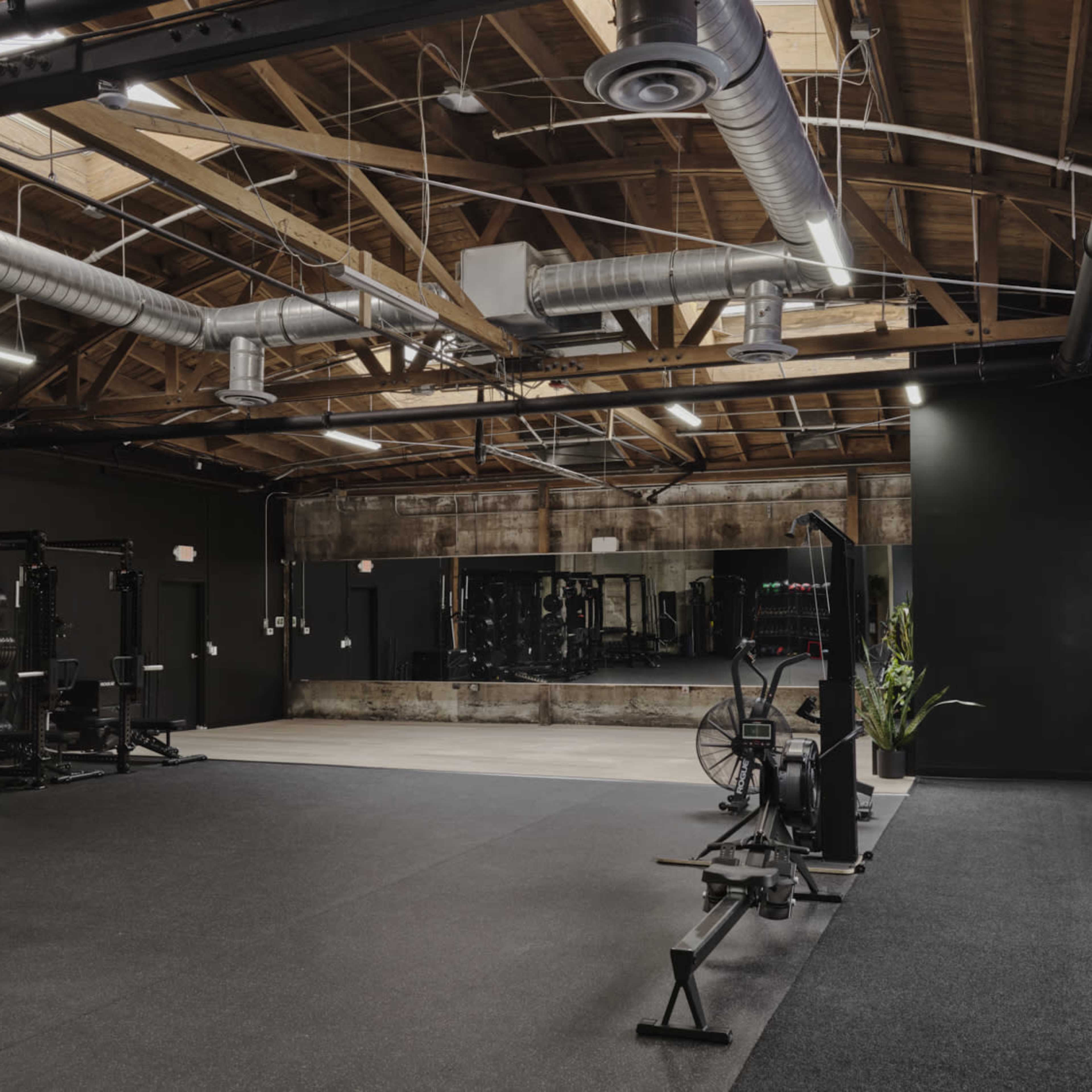 A spacious gym with various workout equipment, including weight racks and rowing machines, set against a backdrop of exposed wooden beams and industrial-style lighting.