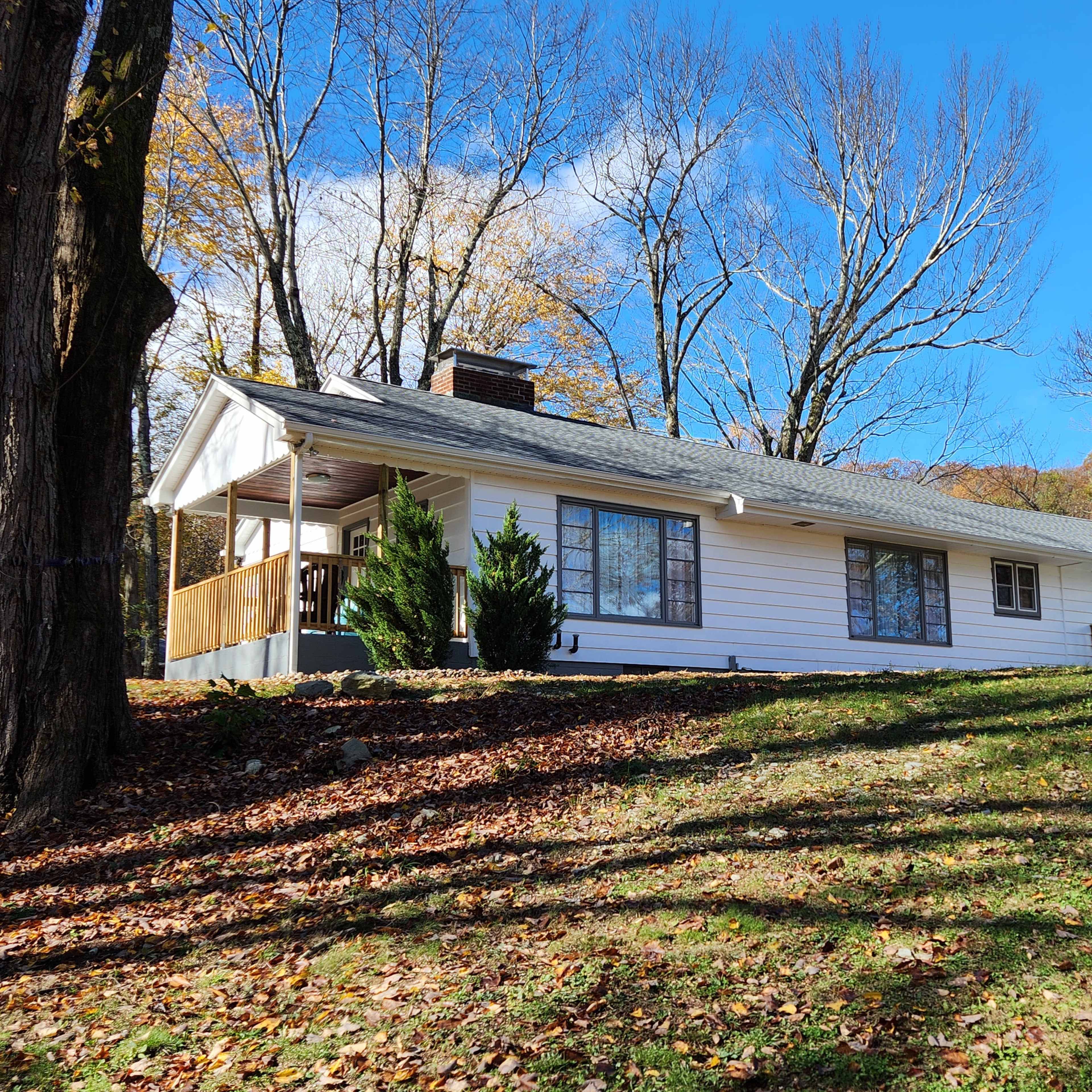 A single-story white house with a gray roof is situated on a hillside, surrounded by bare trees and autumn foliage.