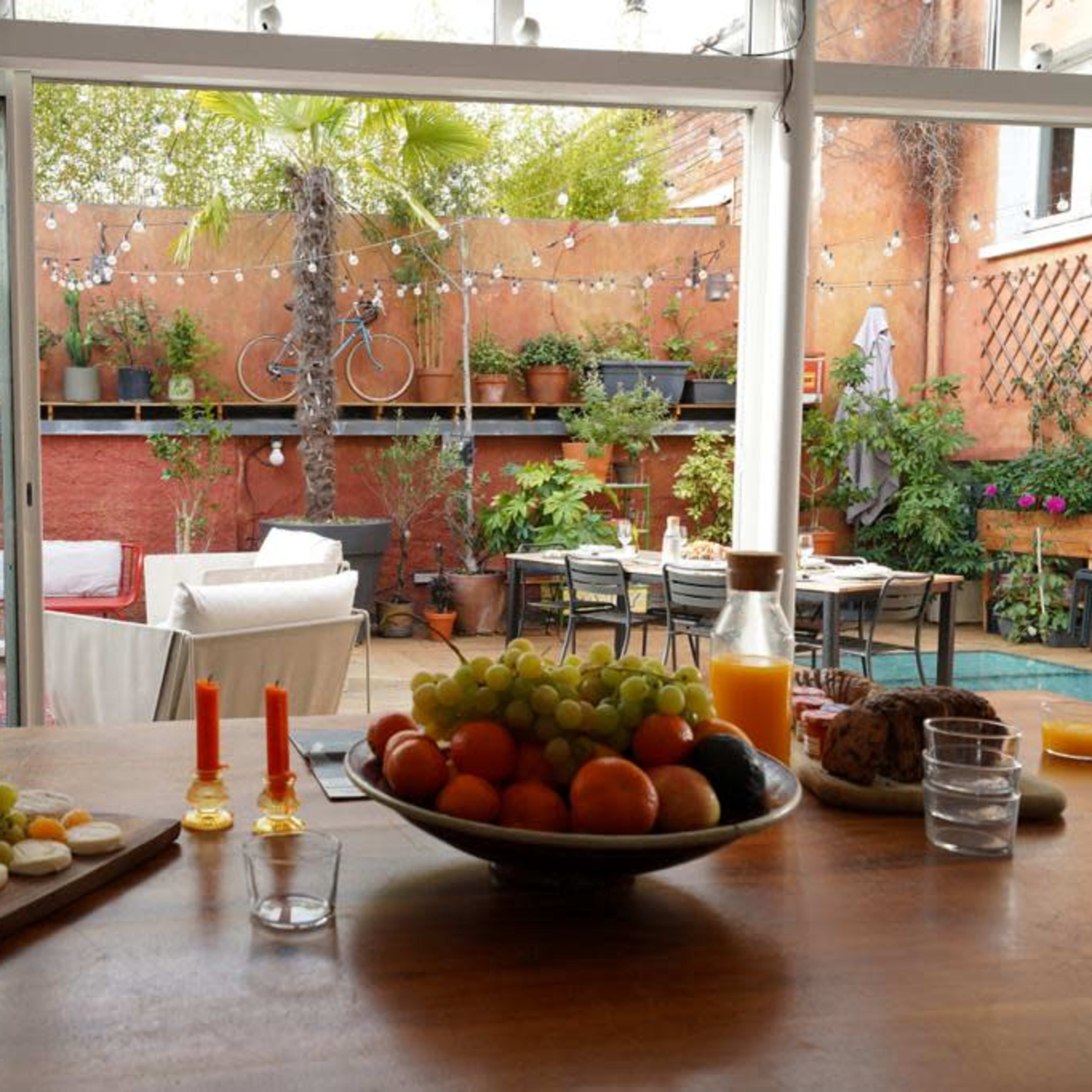 A bright, indoor dining area features a wooden table set with fruits and snacks, overlooking a lush garden with outdoor seating and string lights.