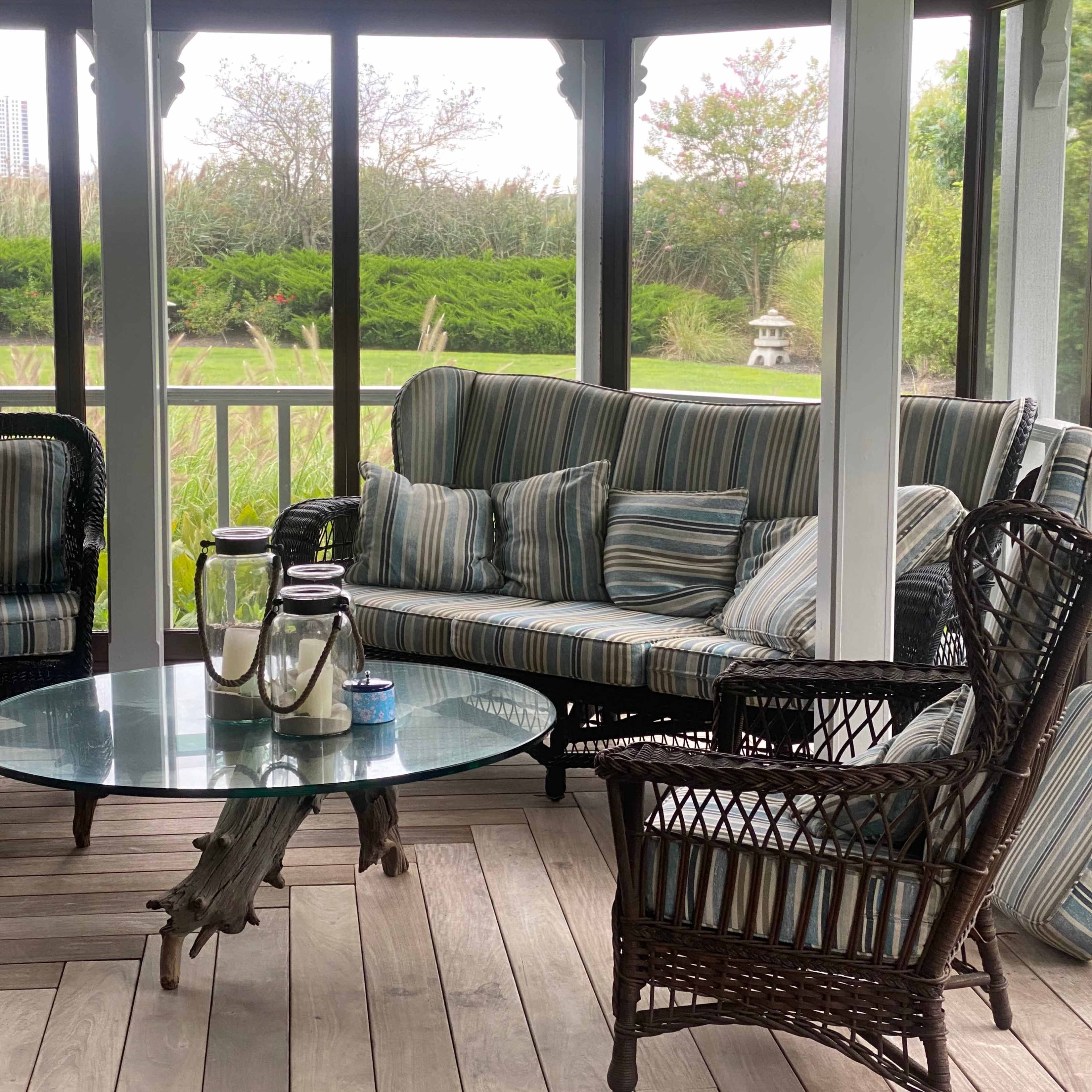 A screened porch with a striped sofa, wicker chairs, and a glass coffee table, overlooking a green landscape.