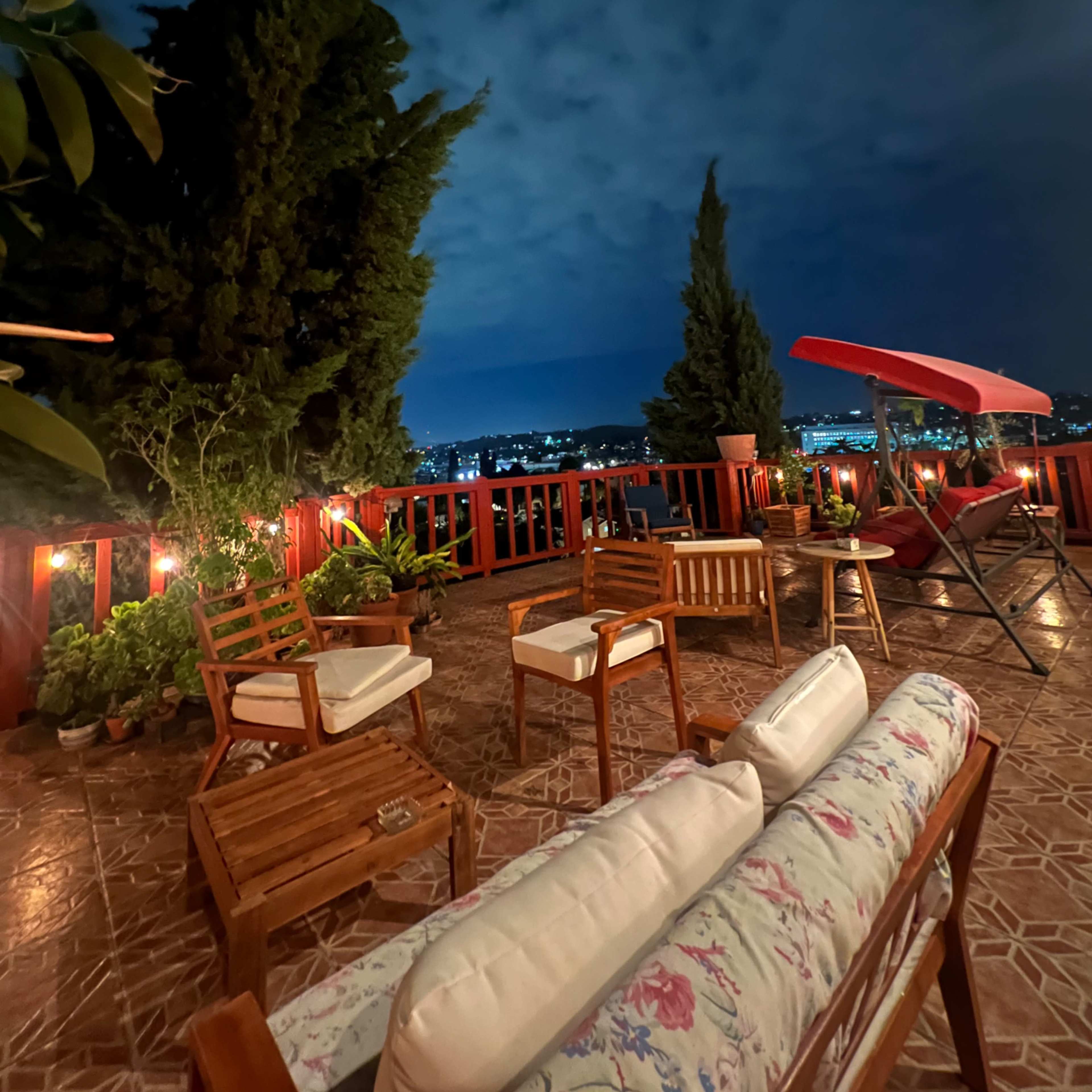 A patio area with wooden furniture and potted plants overlooks a city skyline under a night sky.