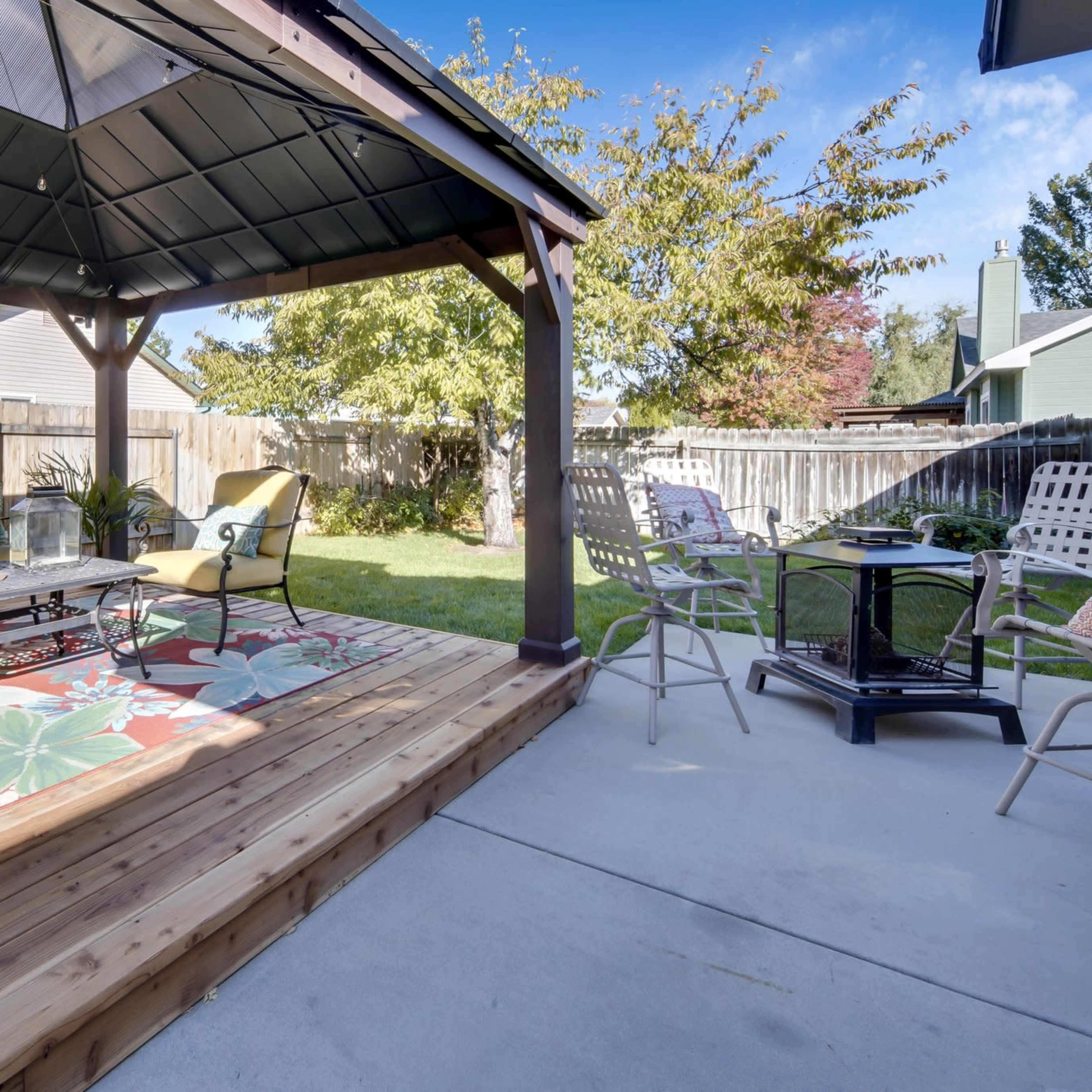 A backyard gazebo with seating and a coffee table on a patterned rug, surrounded by greenery and a wooden fence.