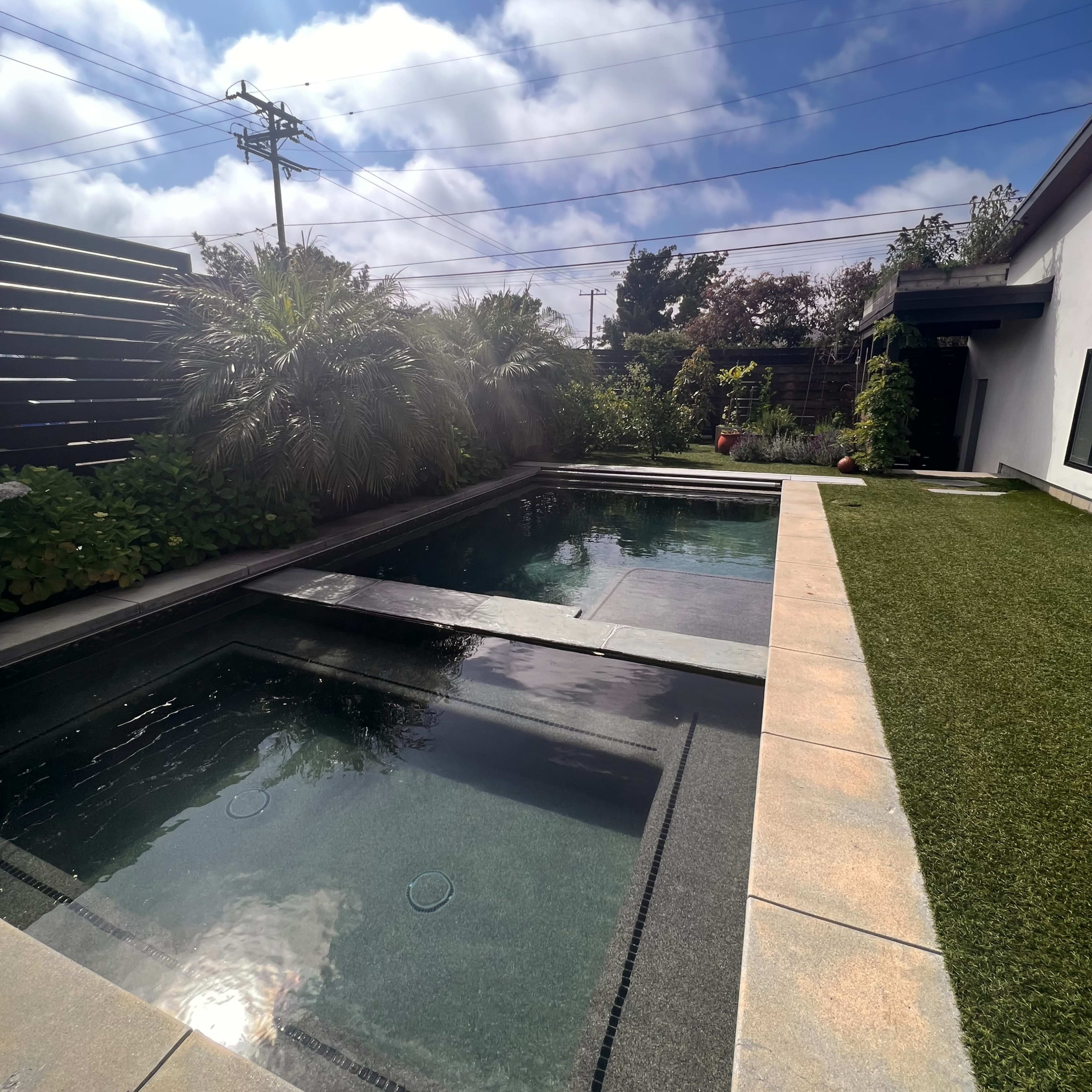 A modern outdoor space featuring two swimming pools surrounded by greenery and a patch of artificial grass under a partly cloudy sky.