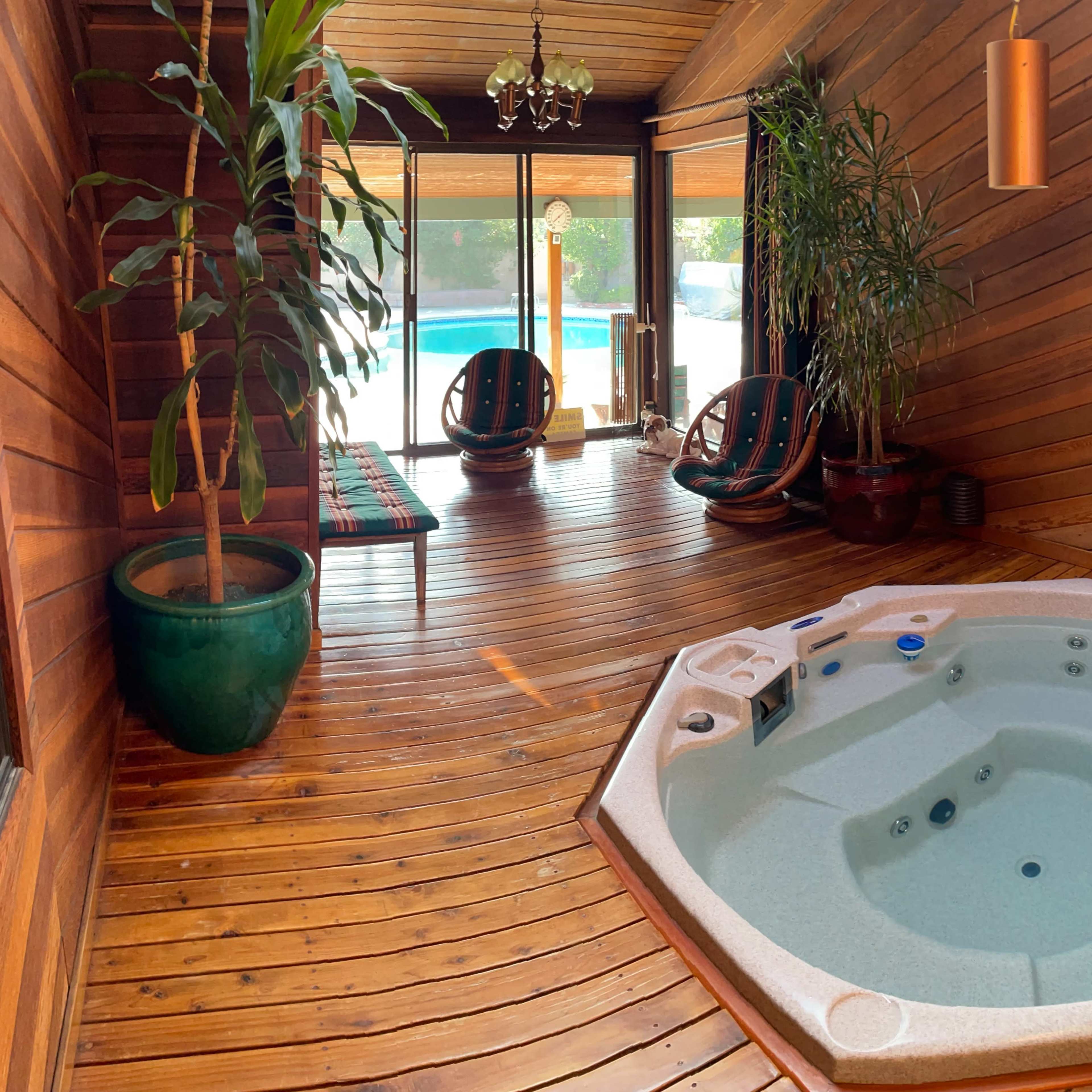 A wooden room featuring a hot tub, large windows, and two chairs beside potted plants.