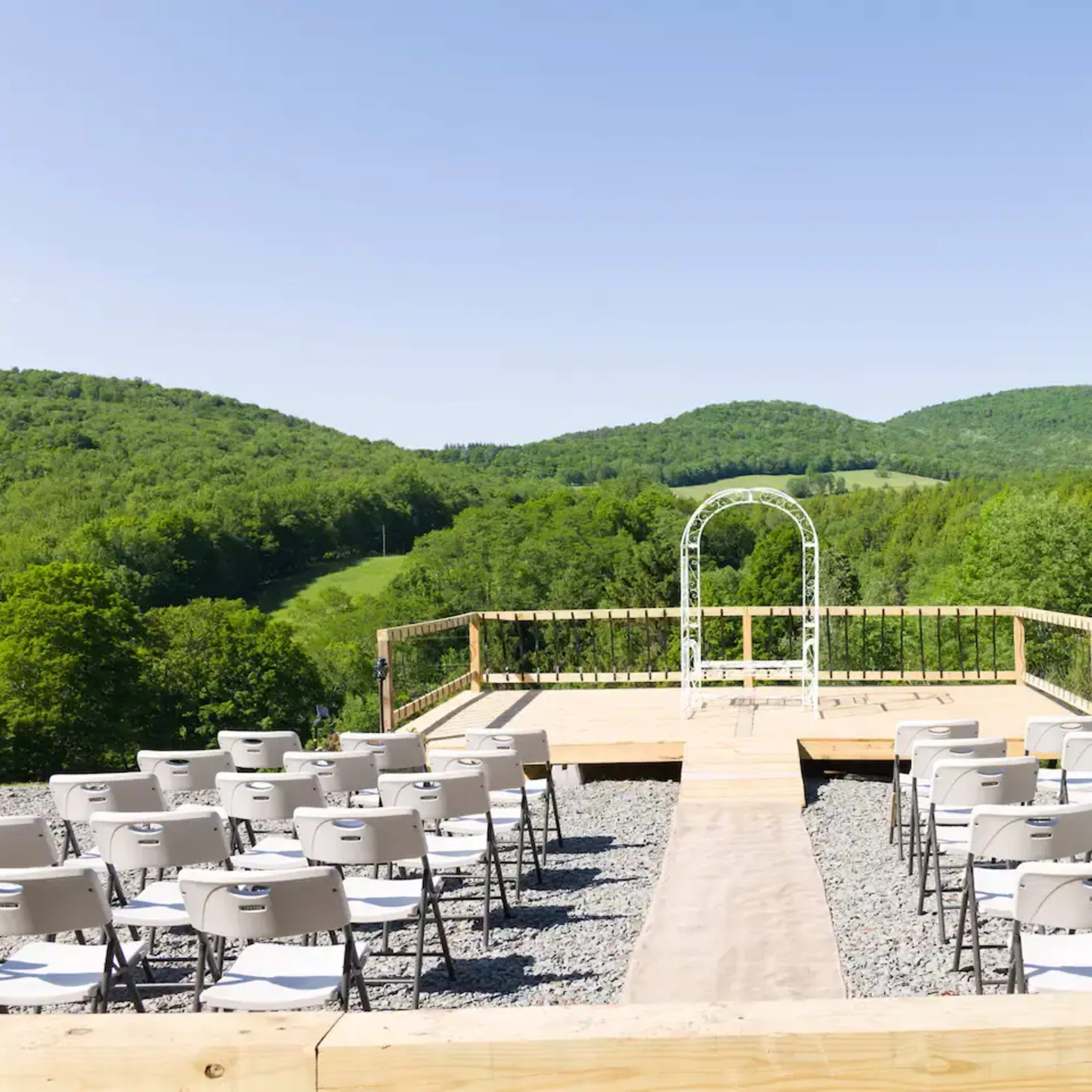 A wooden platform with white chairs arranged for an outdoor wedding ceremony overlooks green hills and a metal arch.