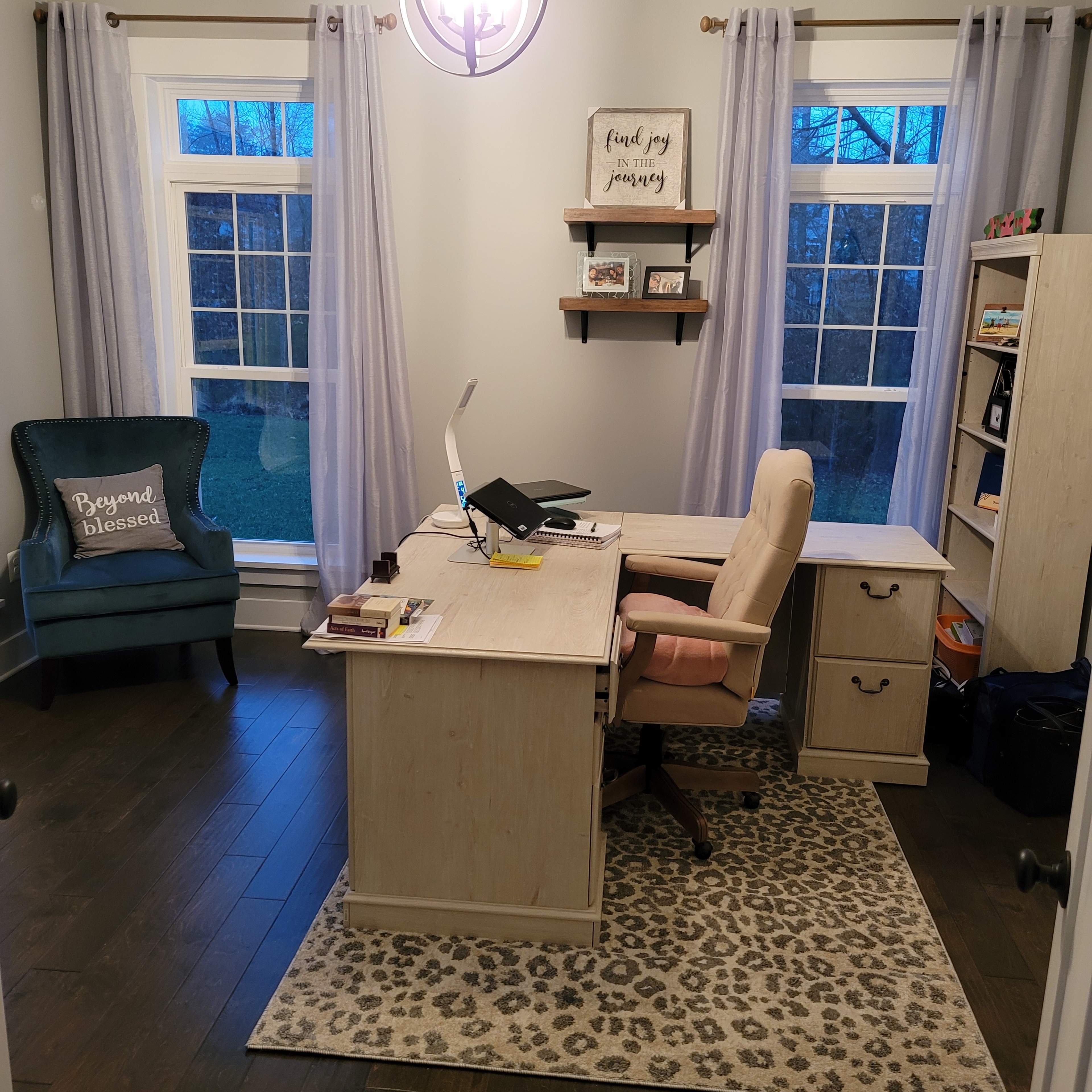 A home office with a desk, a chair, bookshelves, a large window, and a decorative rug.
