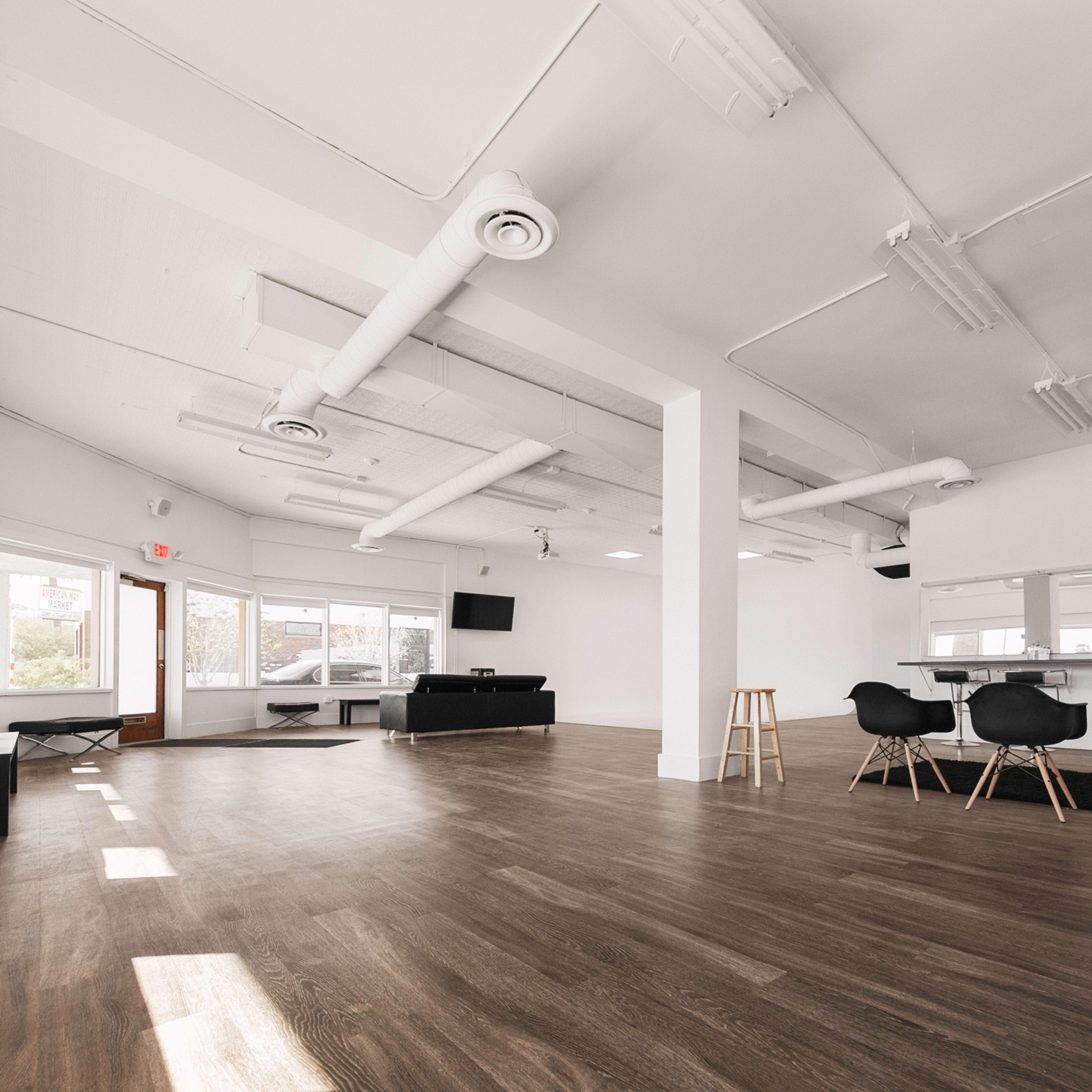 A spacious, bright room with wooden flooring, large windows, and minimalistic furnishings, including a long table and several black chairs.