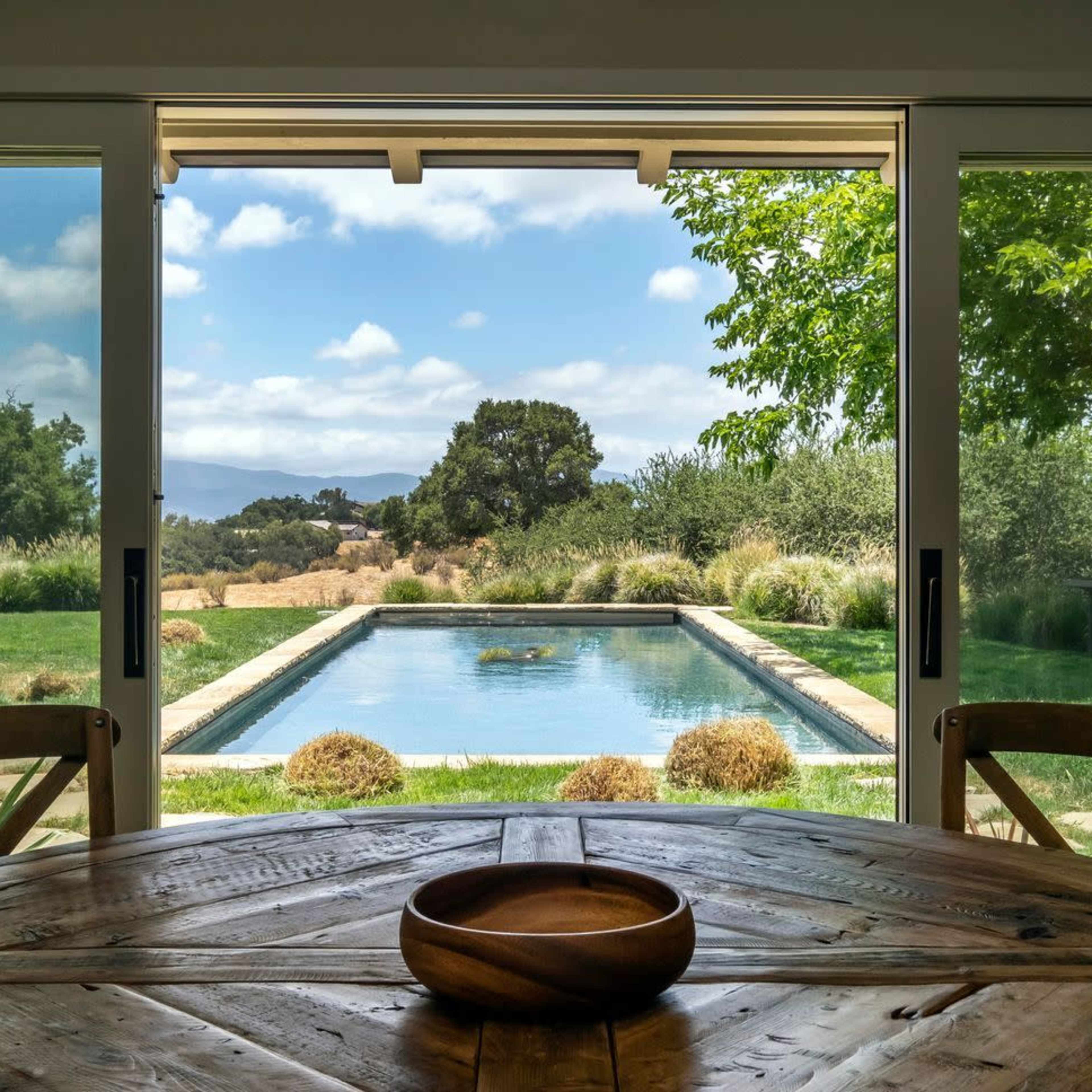 A wooden dining table sits in front of large open doors, revealing a pool and landscaped backyard view with trees and clouds in the sky.